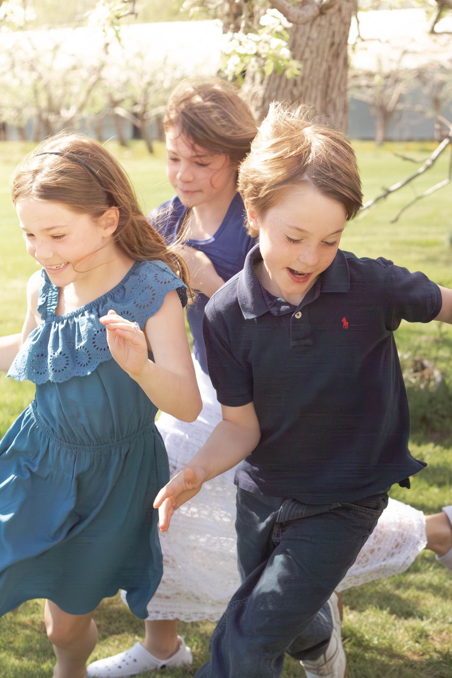 Three children running and playing together outdoors in a sunny park with trees in the background.