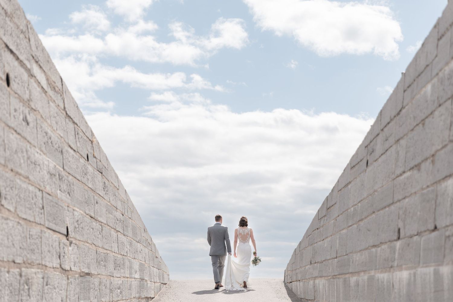 Bride and groom walking between stone walls toward open sky on their wedding day.