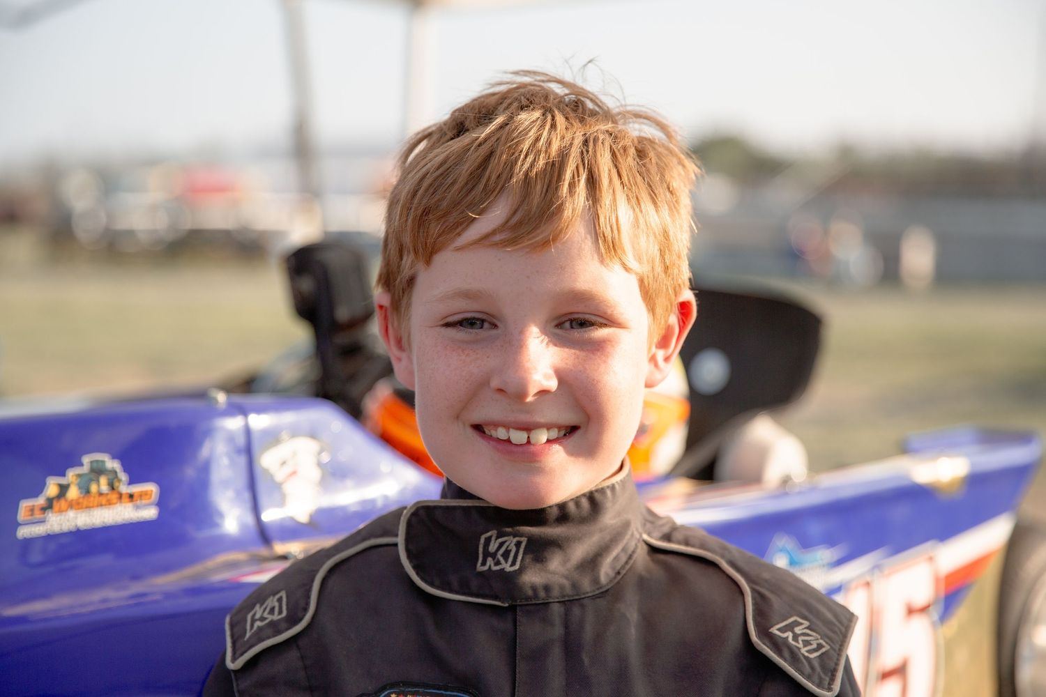 Young go-kart racer wearing a black racing suit stands in front of a blue racing vehicle on a sunny day.
