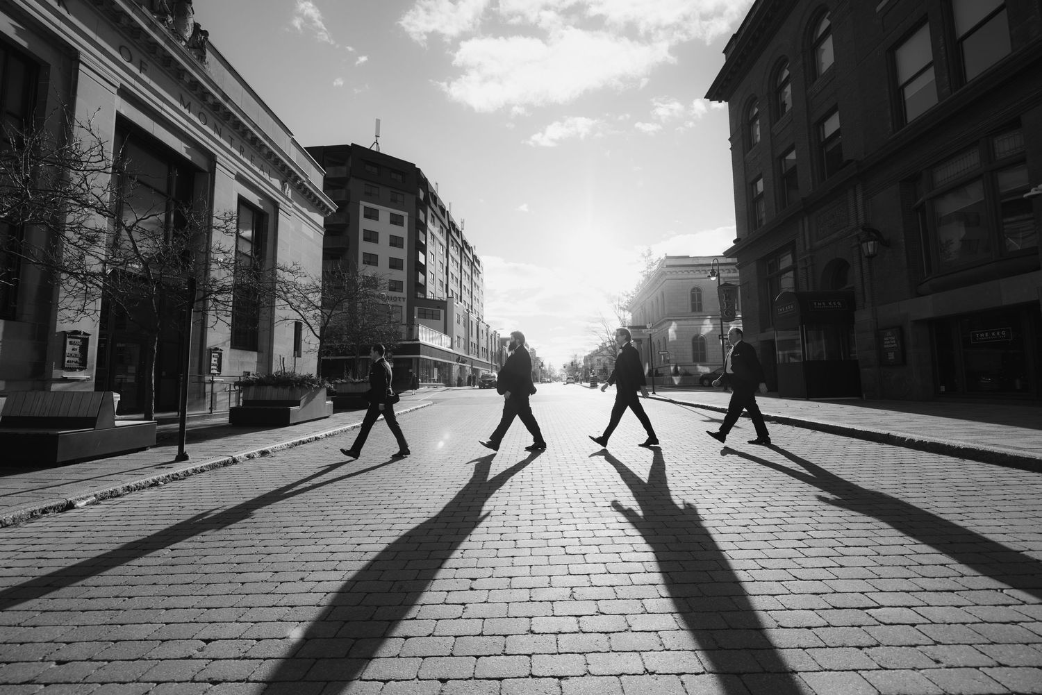 Long shadows cast by pedestrians walking on a brick sidewalk in black and white urban street photography.