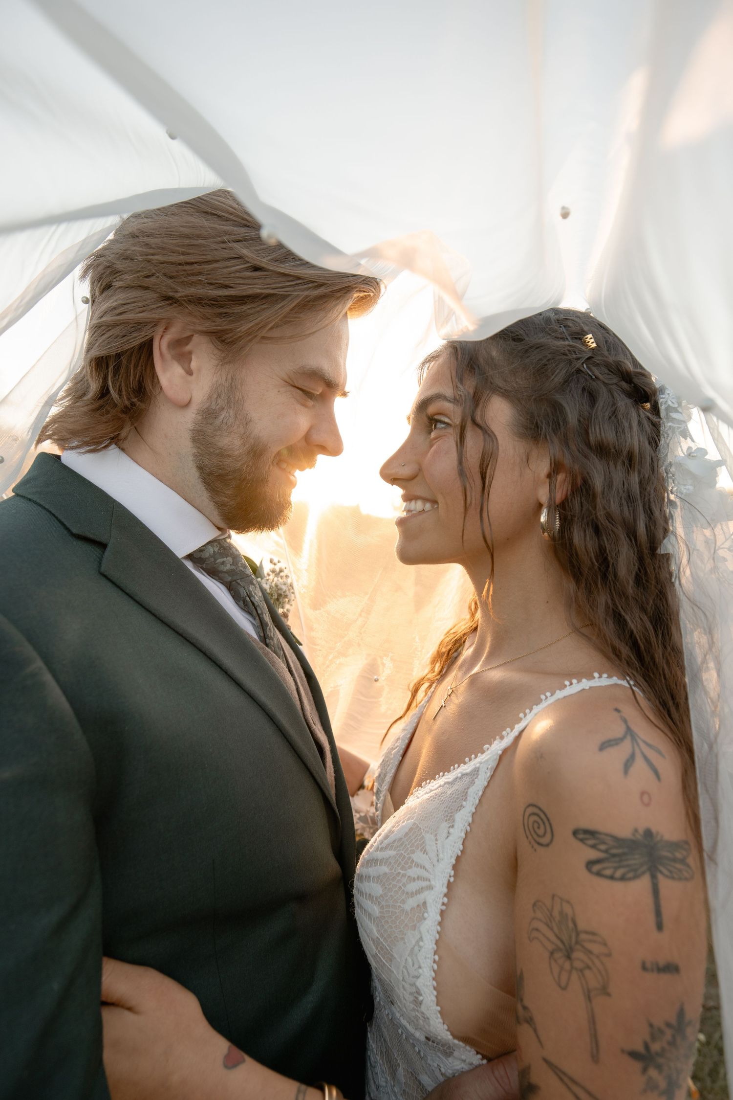 A romantic wedding couple embraces during golden hour sunset with the bride showing delicate tattoos on her shoulder.