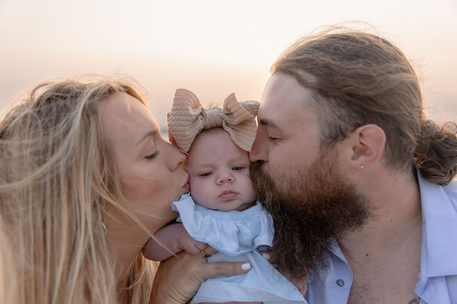 Parents sharing a loving kiss with their baby at sunset by the ocean.