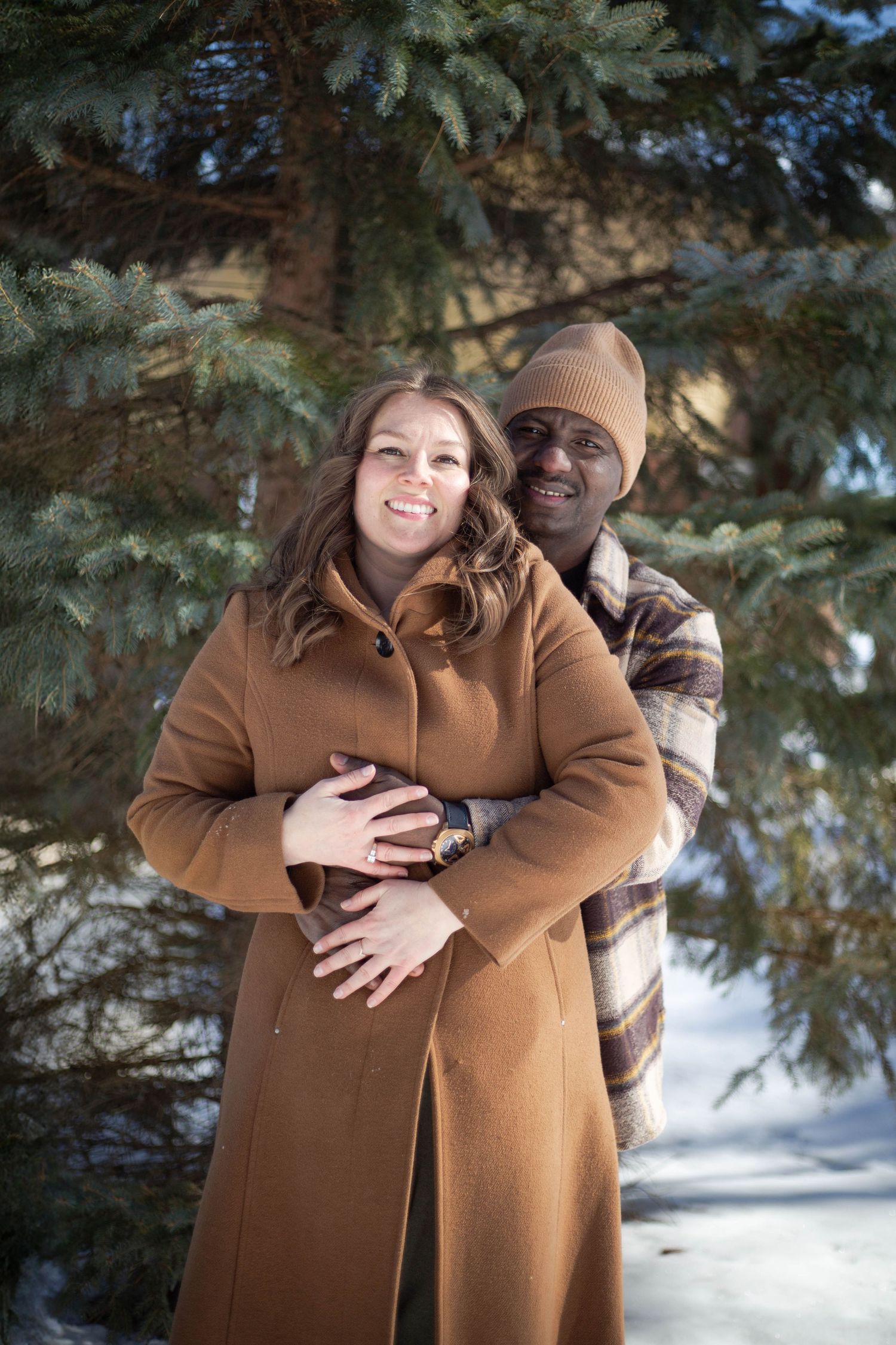 A couple embraces in front of evergreen trees during winter, wearing warm coats and beanies.