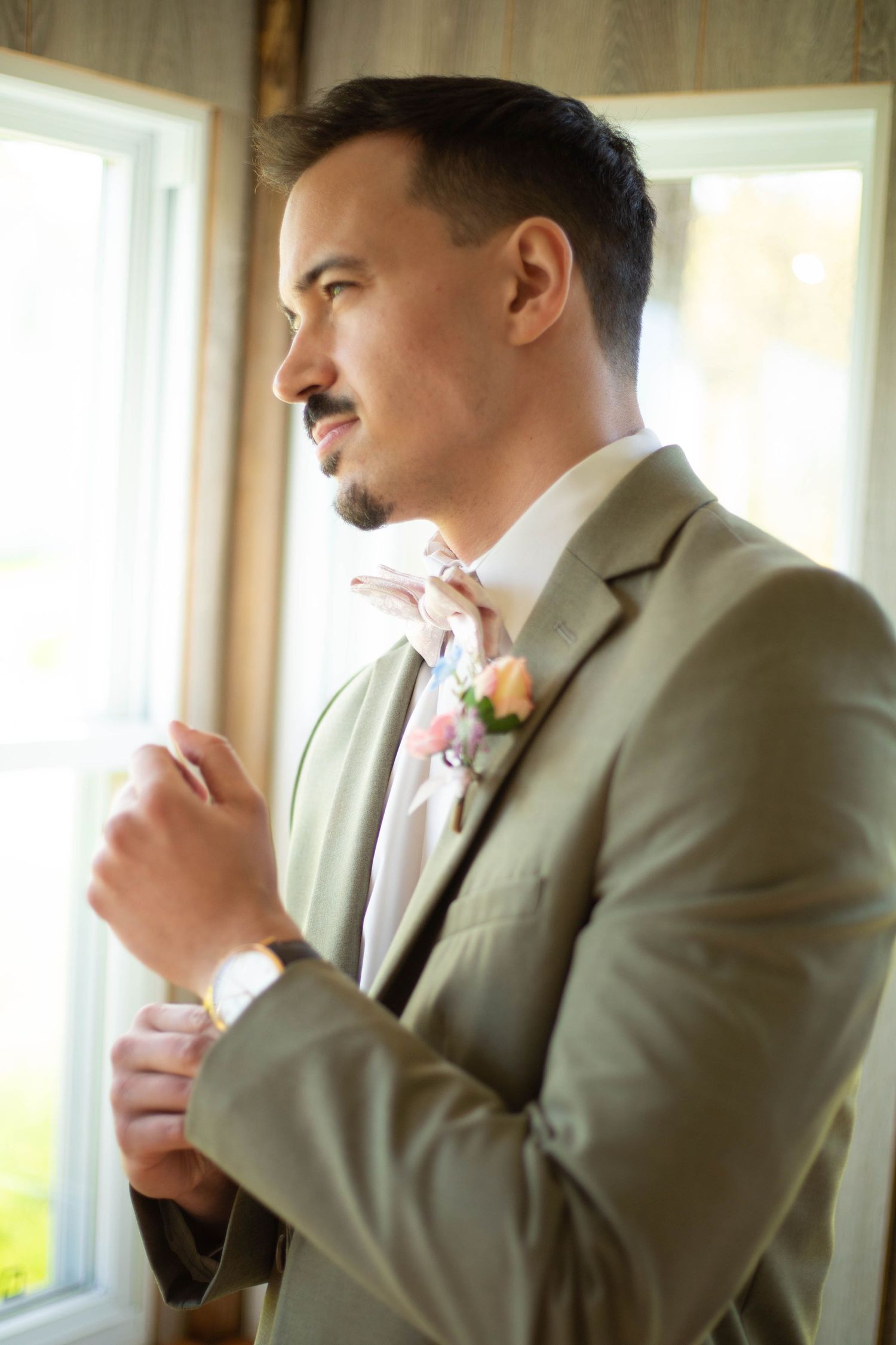 A person in a beige suit adjusts their tie while looking out a sunlit window.