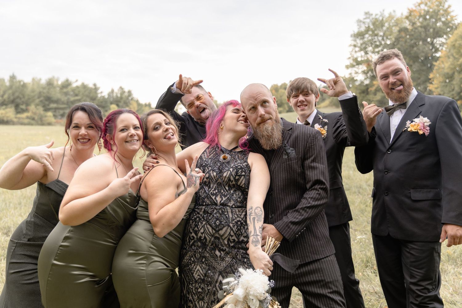 Wedding party in formal attire poses together outdoors making silly faces and gestures on a sunny day.