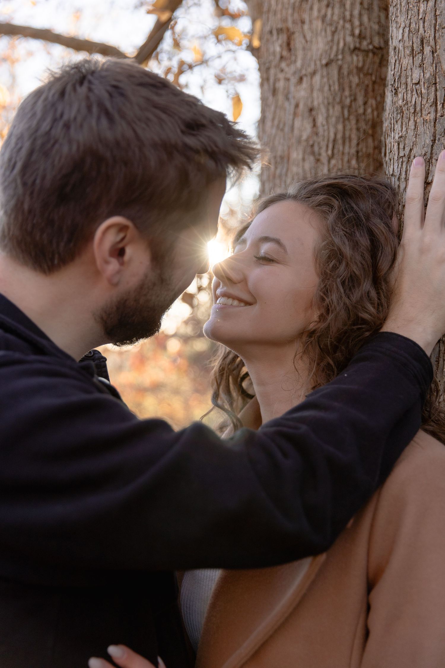 A romantic couple shares an intimate moment against a tree trunk at sunset with warm golden lighting.