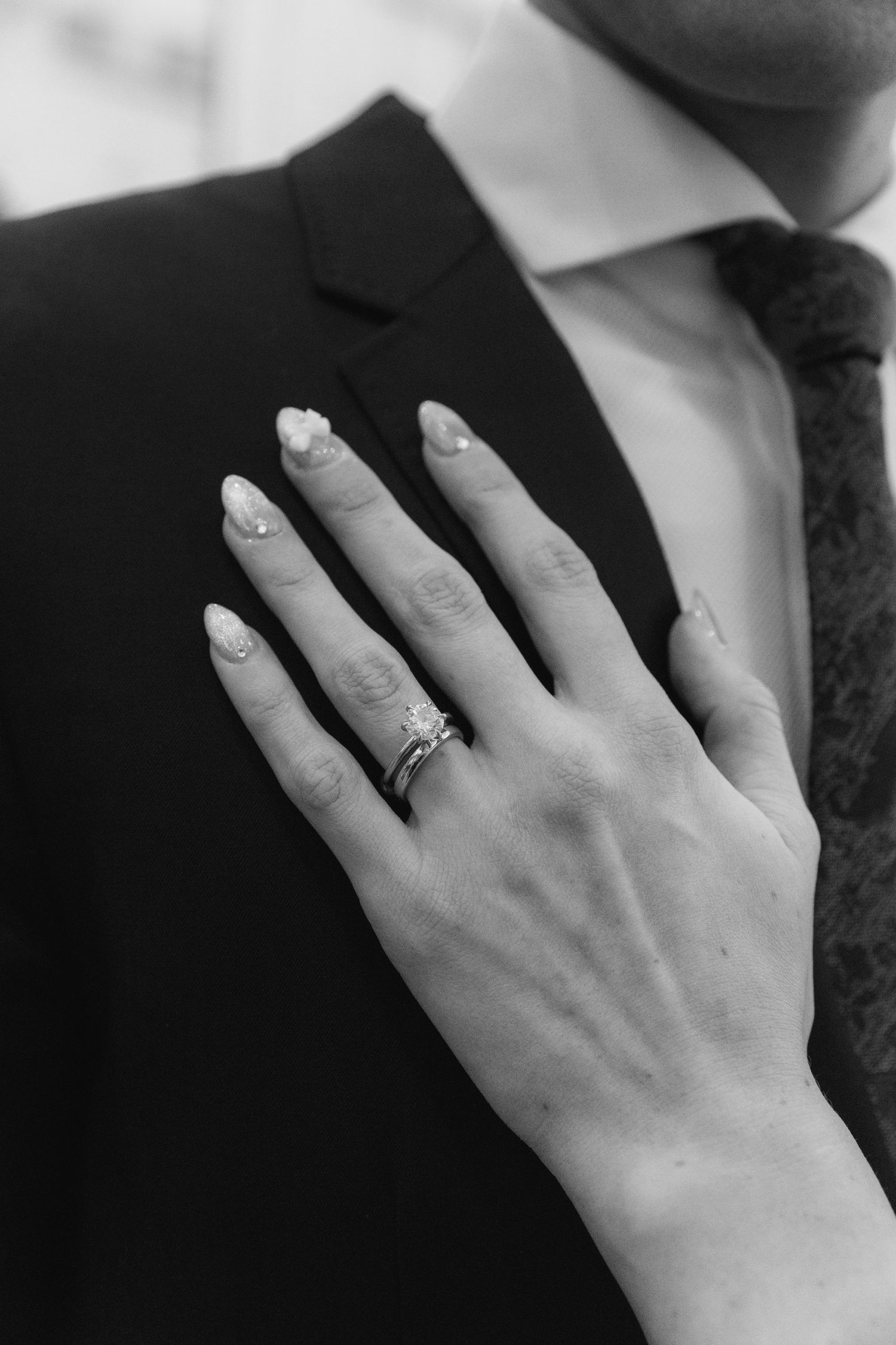 Close-up of a hand wearing an elegant engagement ring resting on a black suit jacket.