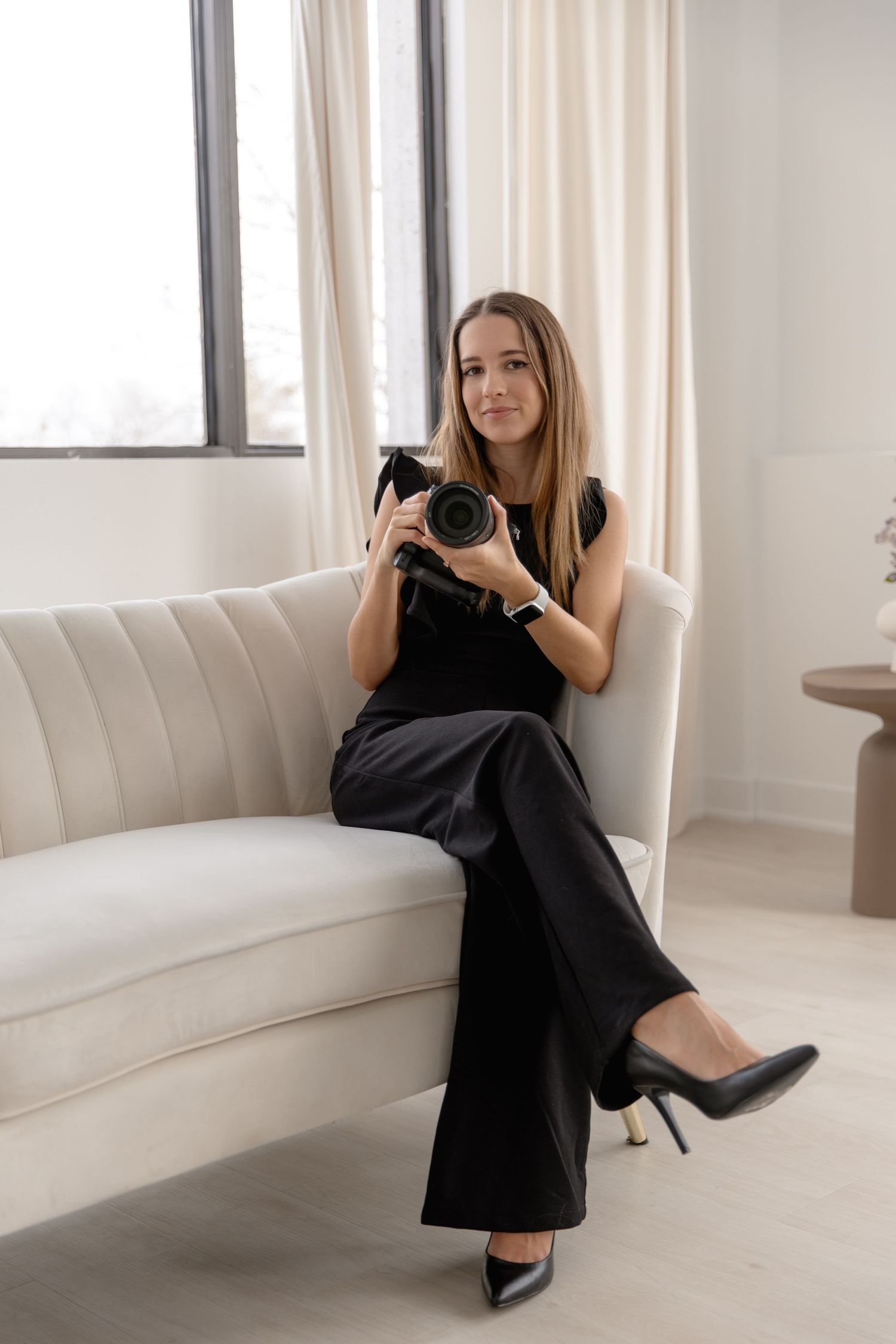 Professional photographer in elegant black outfit sits on white couch holding DSLR camera in bright modern studio setting.