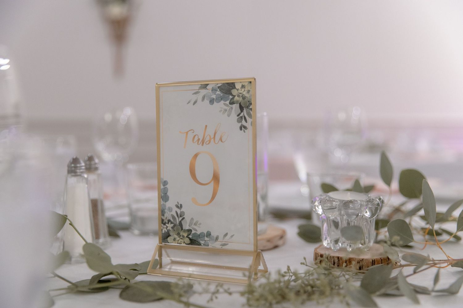 Elegant wedding reception table settings with gold accents, white flowers, and greenery in a soft, romantic atmosphere.