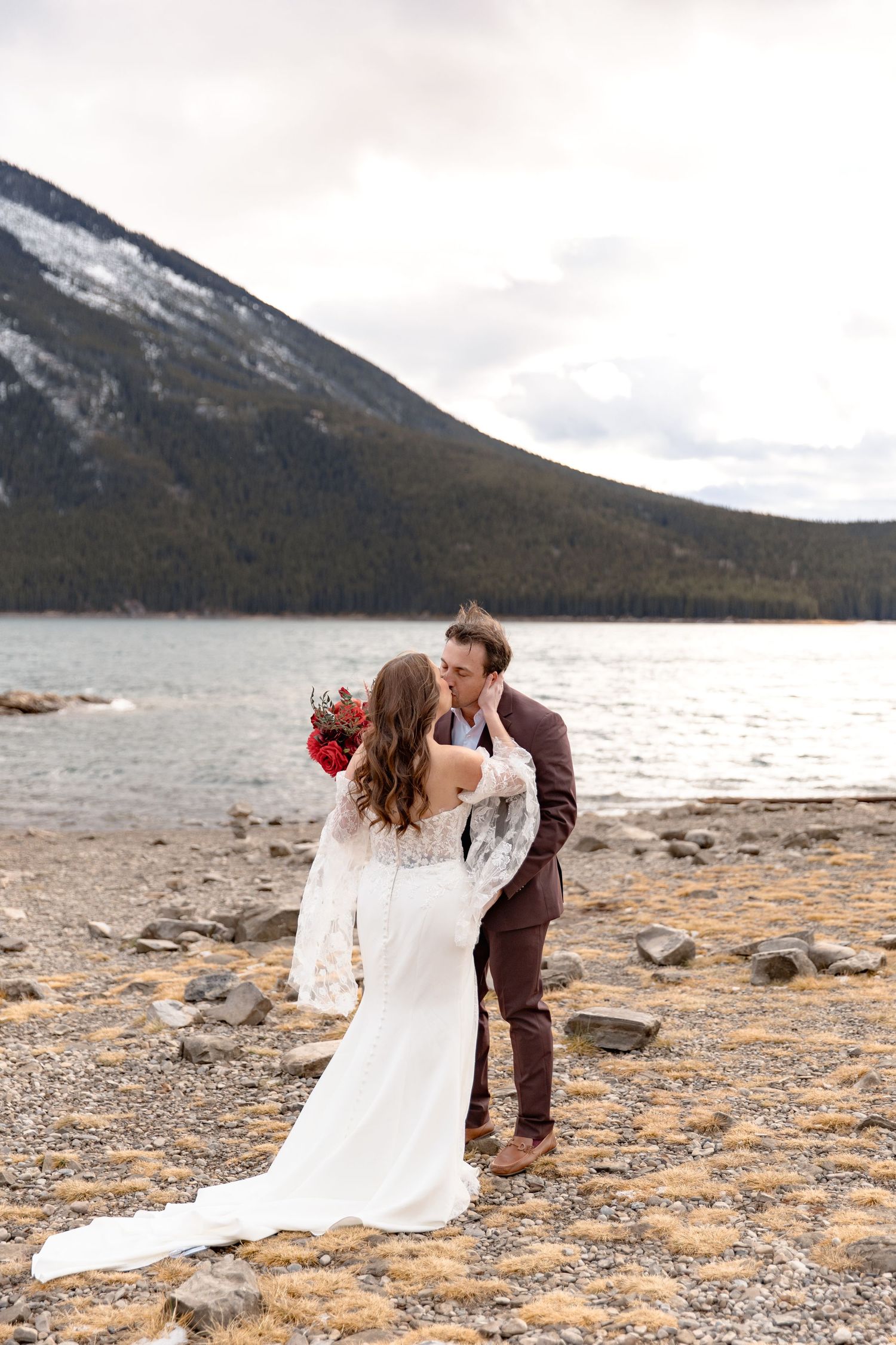 A couple shares a romantic moment on a rocky mountain lake shore in their wedding attire.