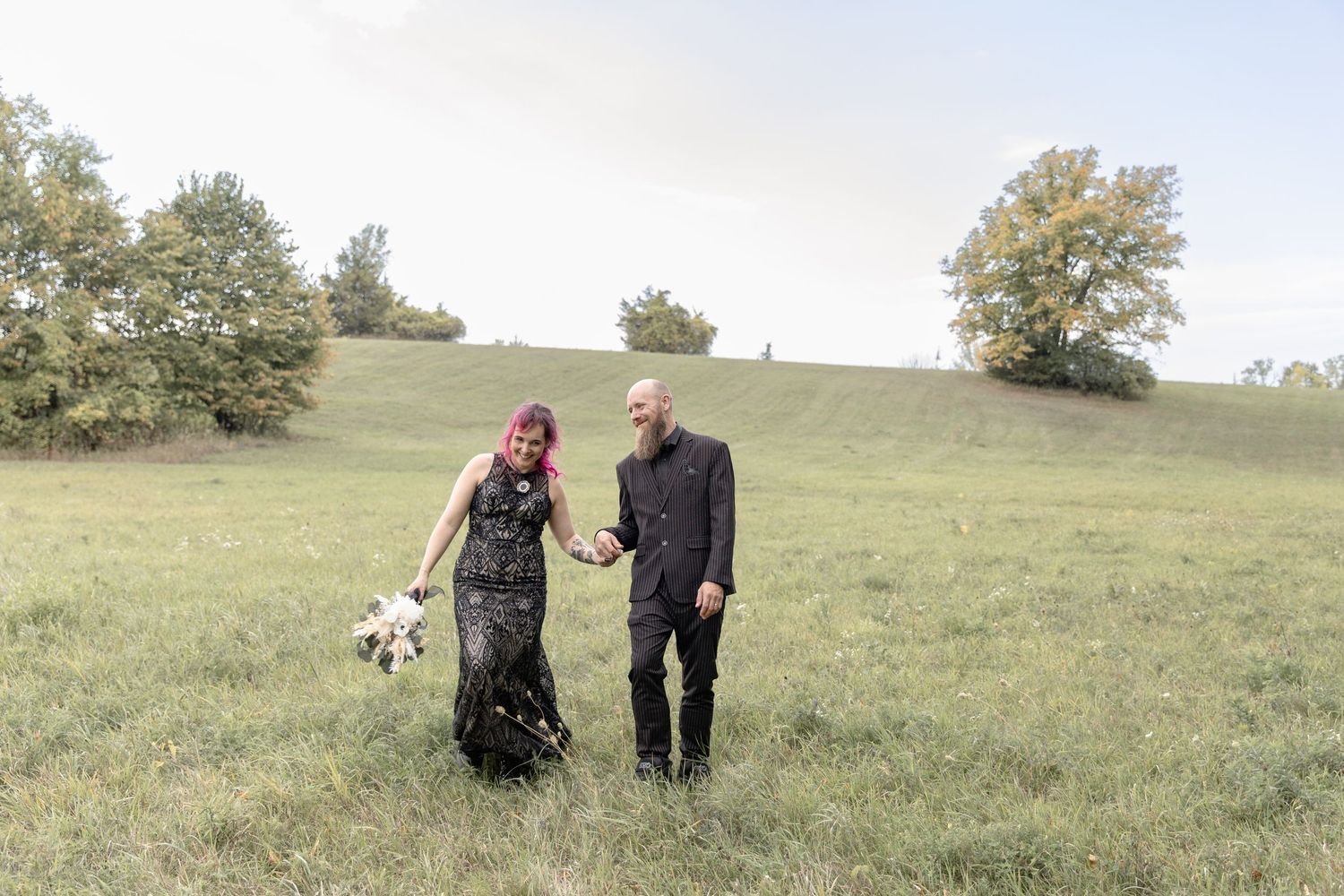 A couple in formal attire stands together in a grassy field with trees on a hillside in the background.