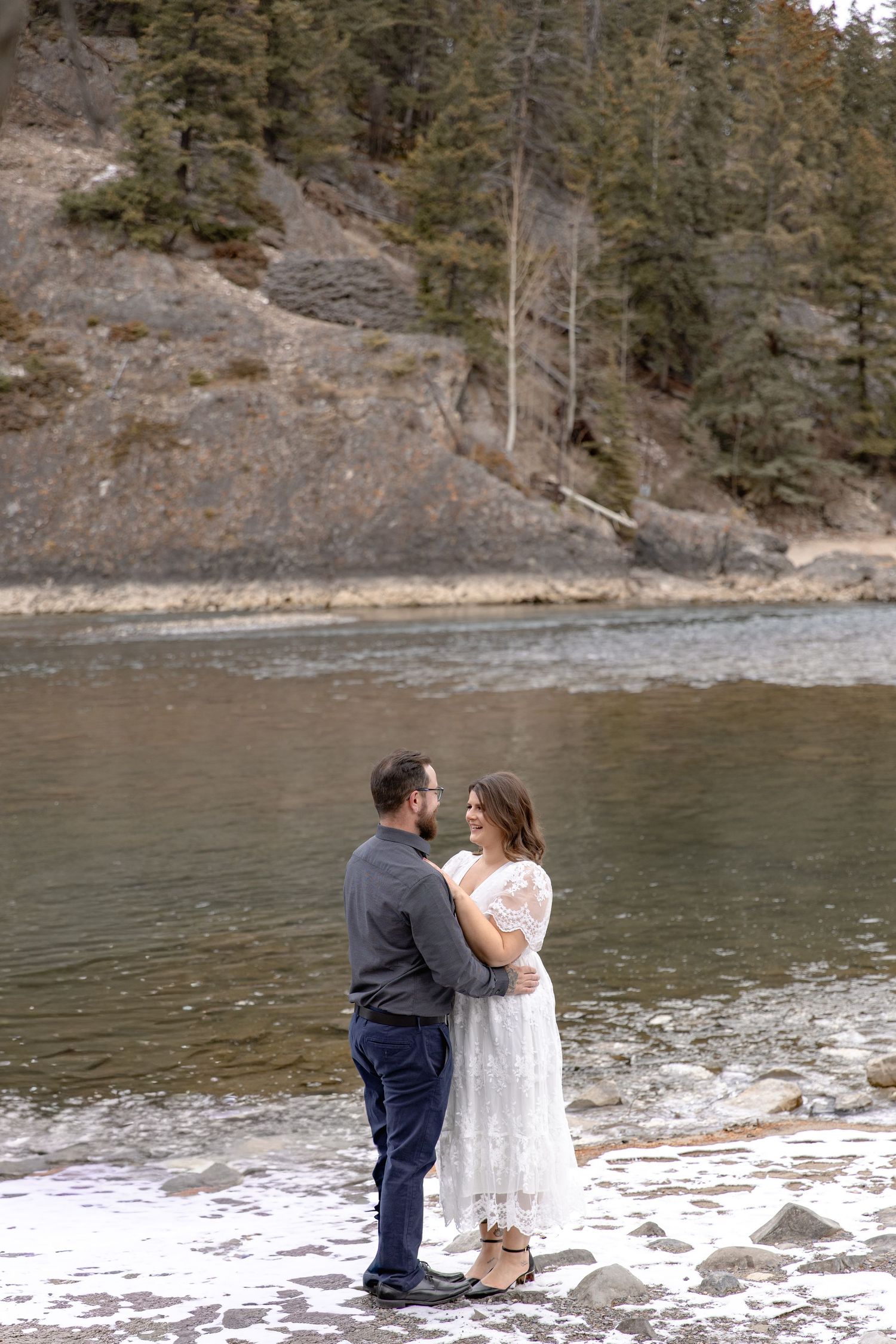 A couple shares an intimate moment on a snowy riverbank surrounded by evergreen trees and rocky slopes.