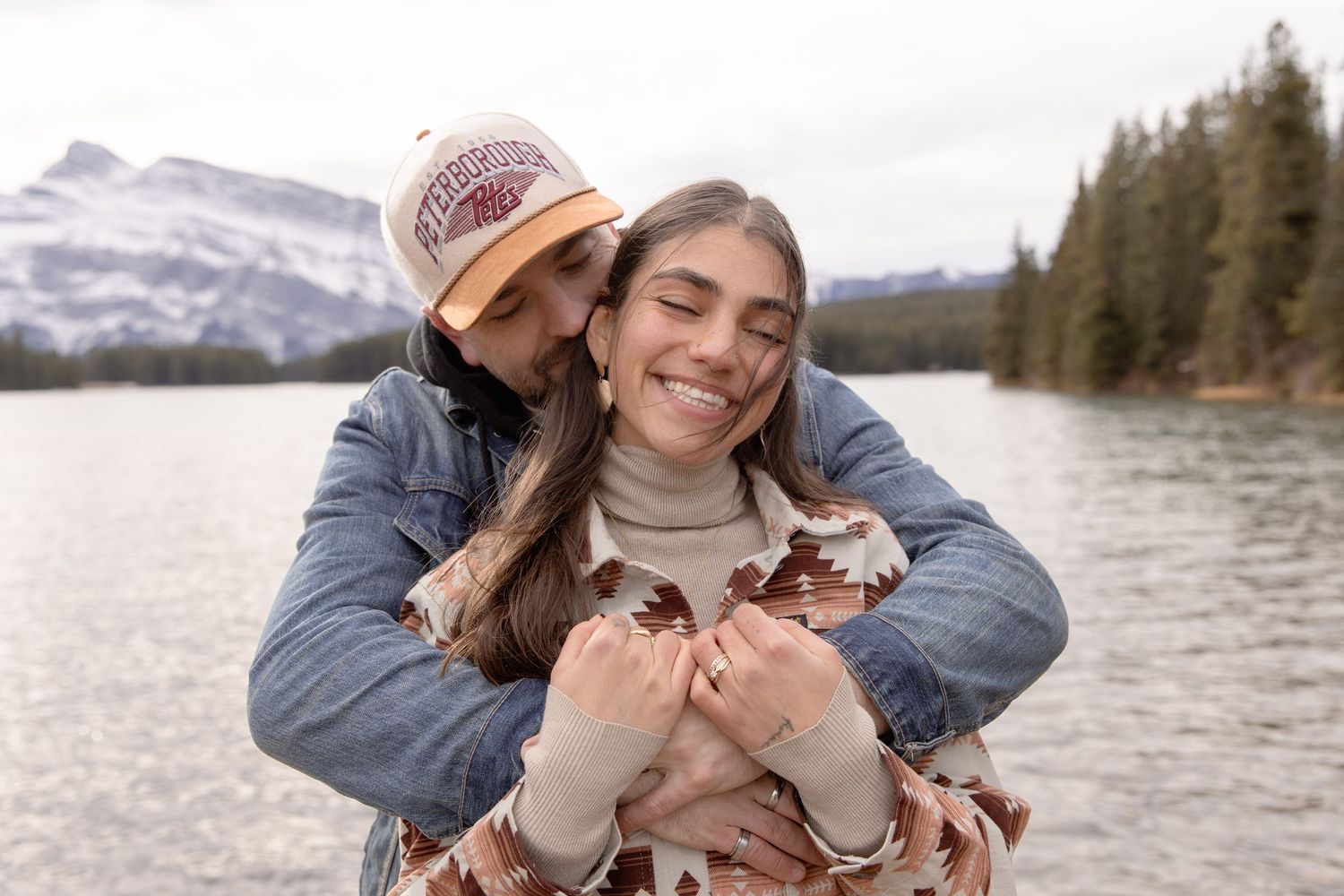 A couple embraces near a scenic mountain lake with snow-capped peaks in the background.