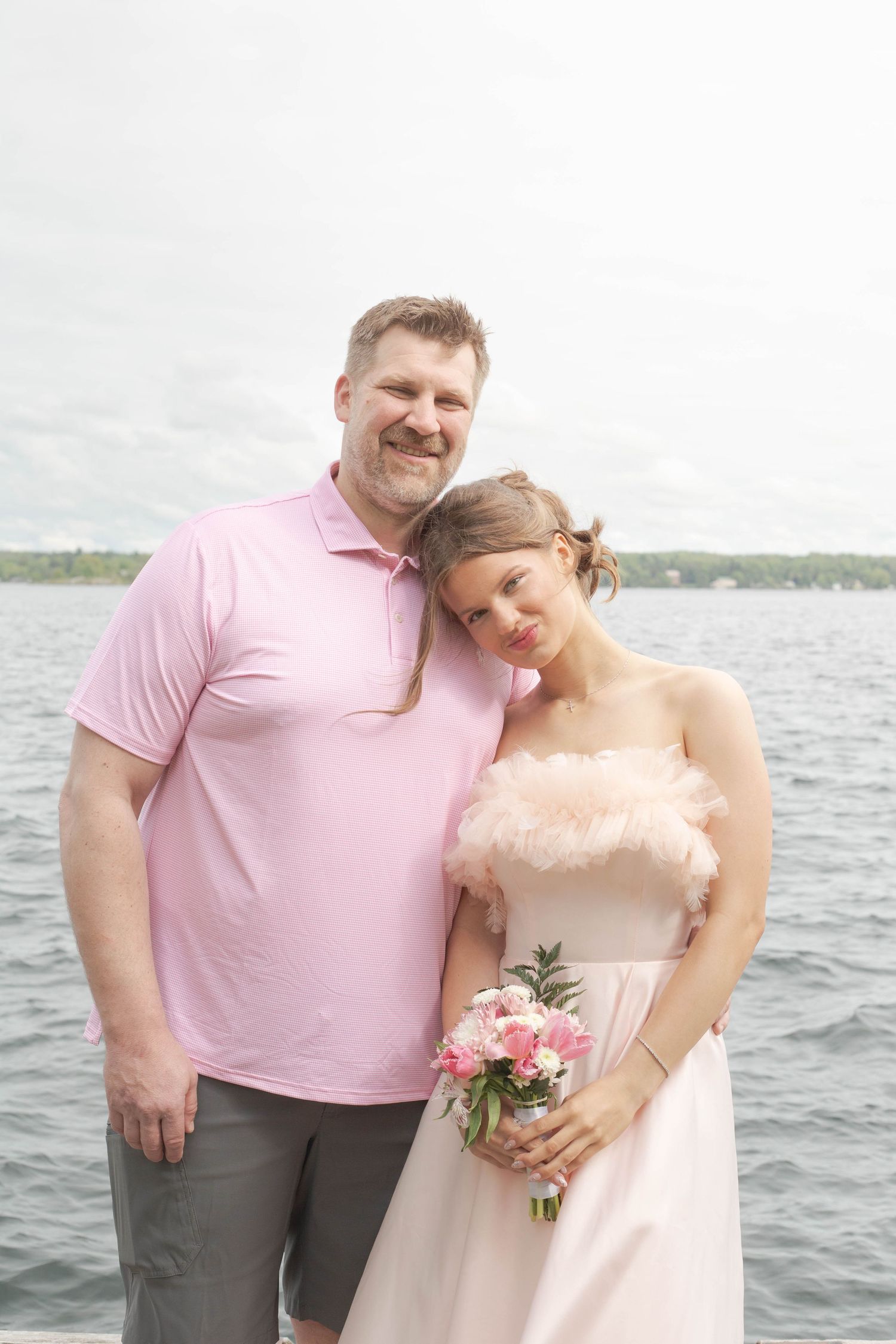 A couple poses by a lake, with one wearing a pink polo shirt and the other in a light blush-colored dress holding flowers.