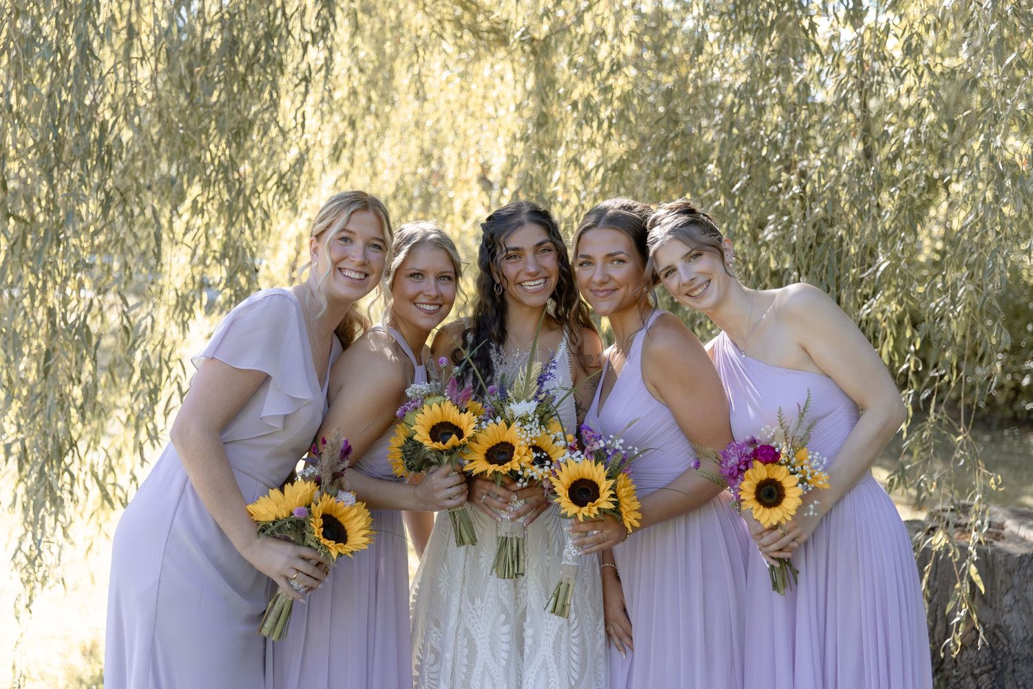 Wedding party wearing light pink dresses holds bright sunflower bouquets while posing under willow trees.