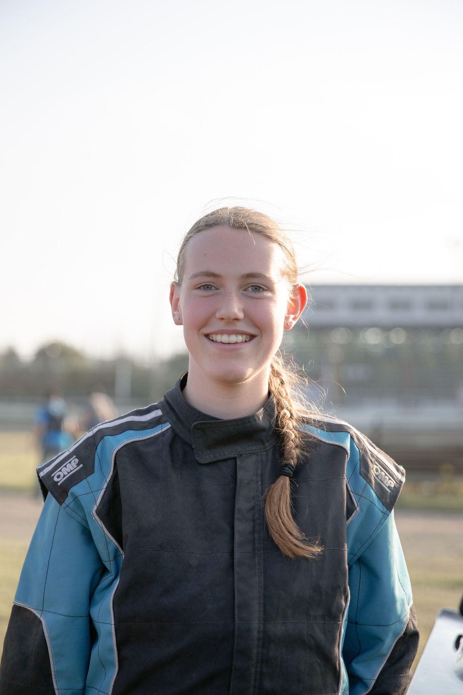 A person wearing a blue jacket and black vest smiles while standing outdoors on a sunny day.