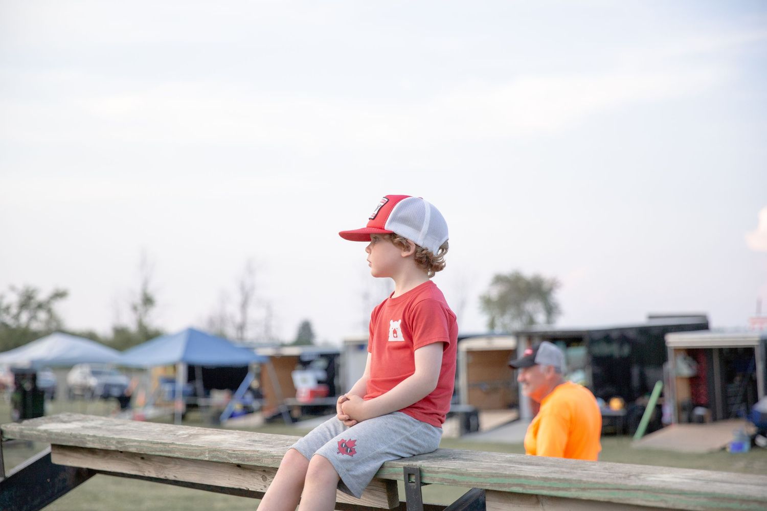 Person in red jacket and grey hat stands outdoors near canopies and vendor setups at a market or event.