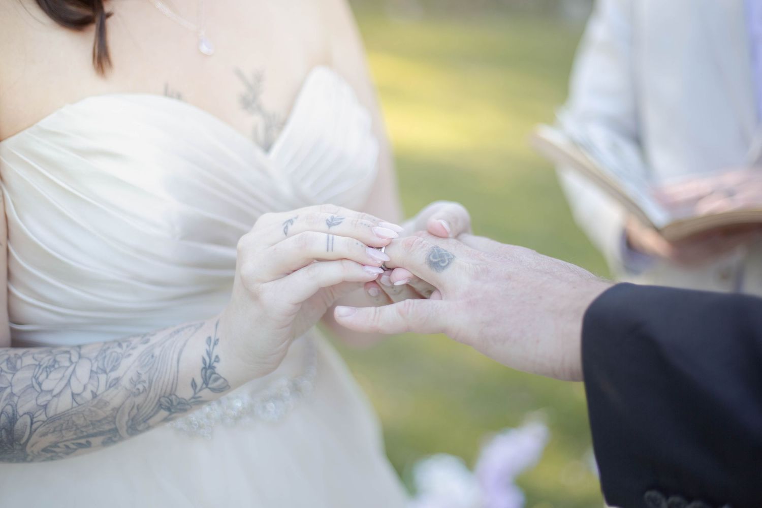 A wedding ceremony moment showing hands exchanging rings with a bride in white dress with tattoos.