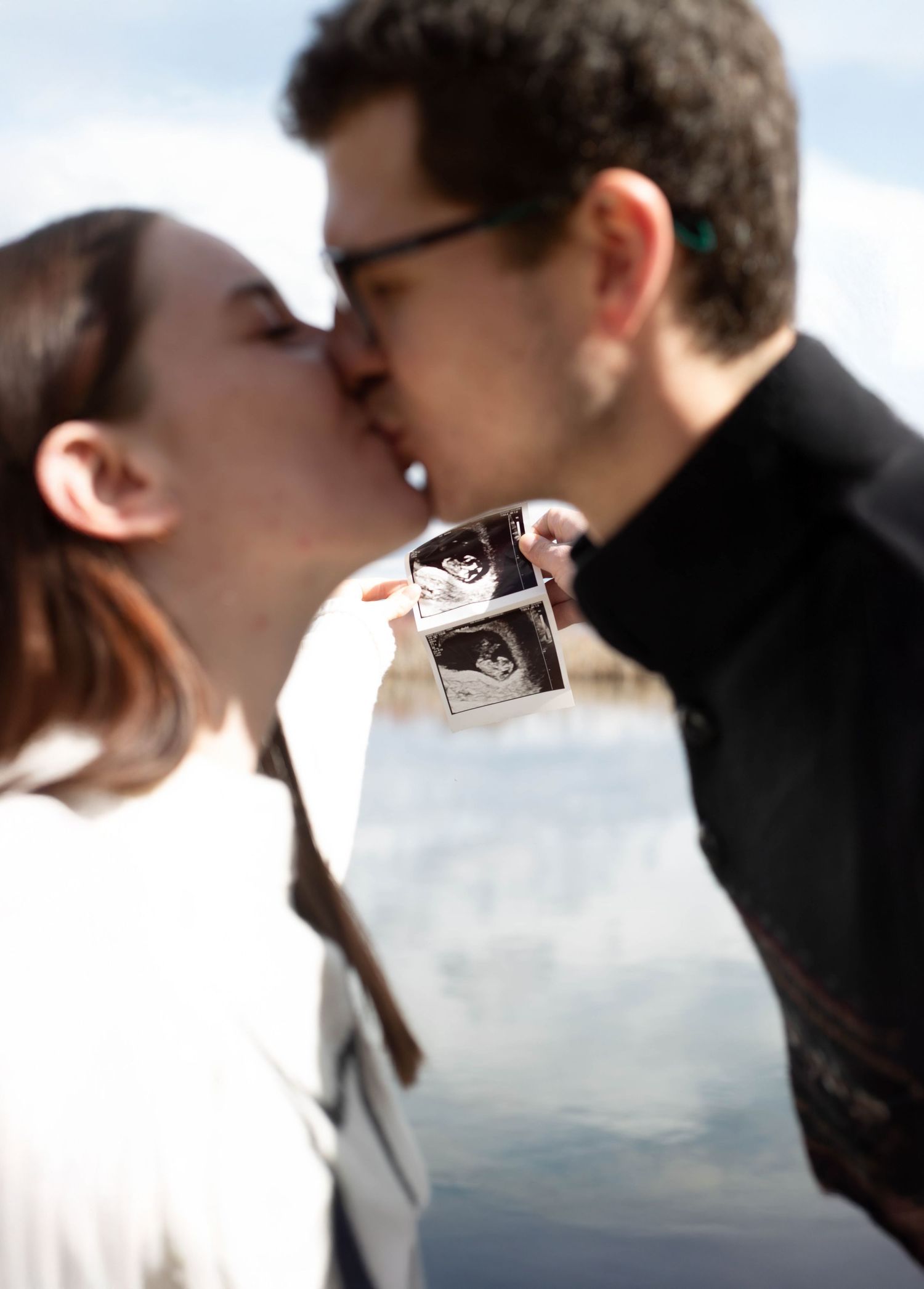 A kissing couple holds up a sonogram photo, announcing their pregnancy against a bright sky.