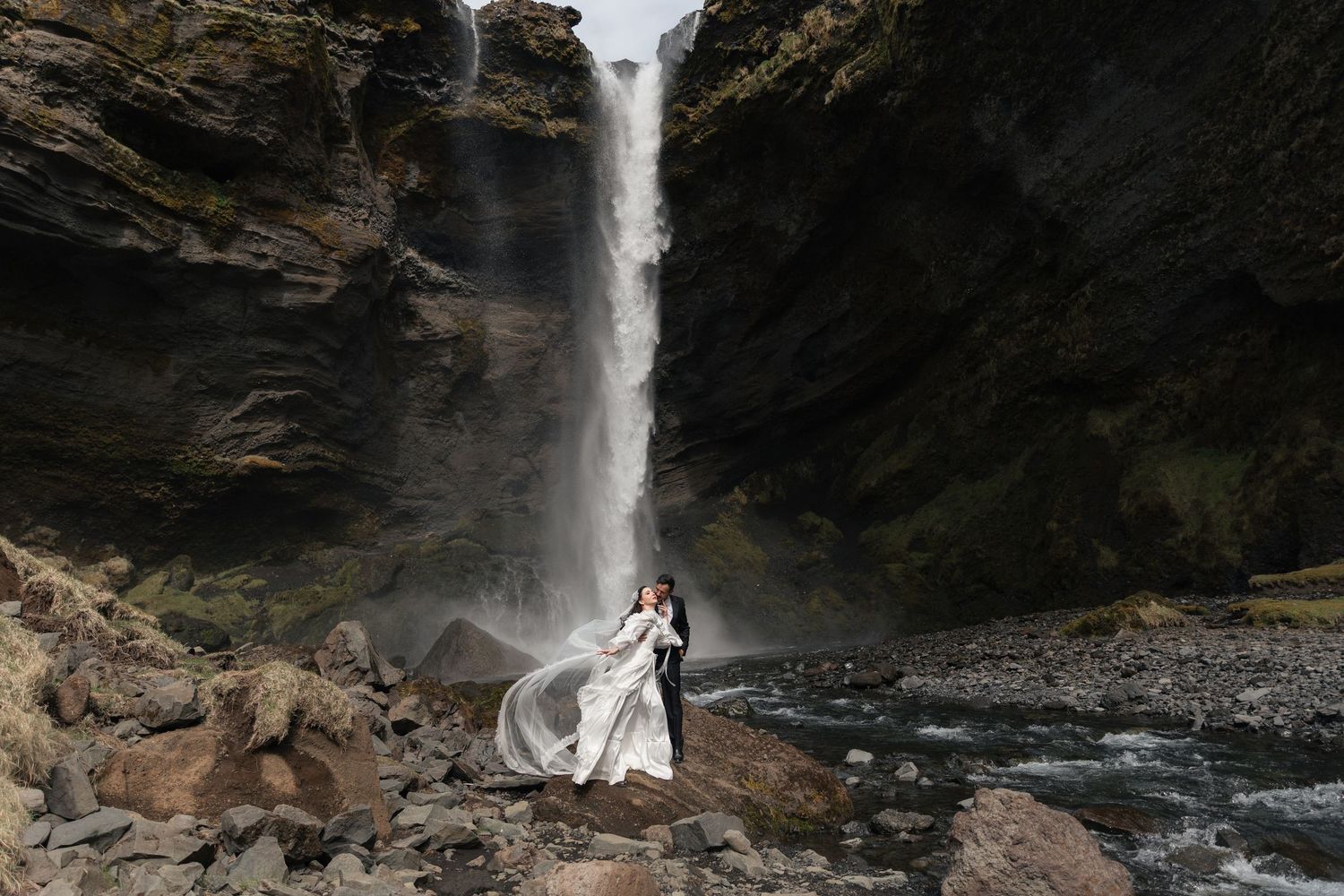 Couple embracing in wedding attire before a dramatic waterfall cascade on rocky terrain.