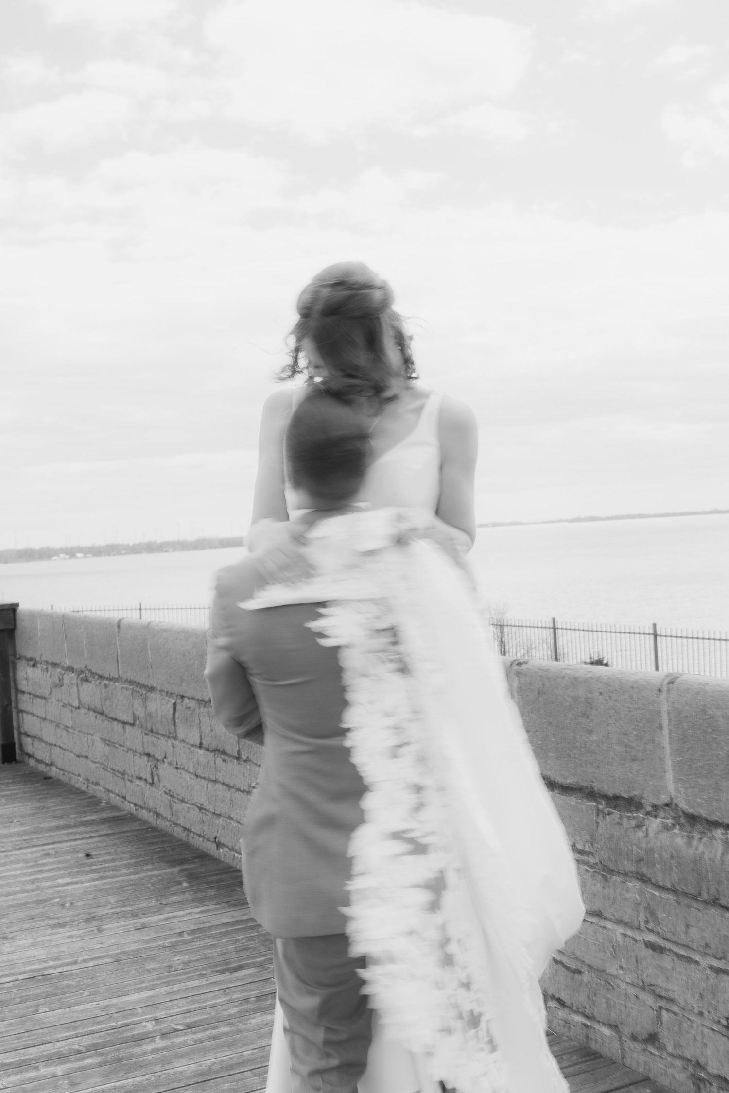 Black and white photo of a bride and groom walking on a waterfront boardwalk, viewed from behind.