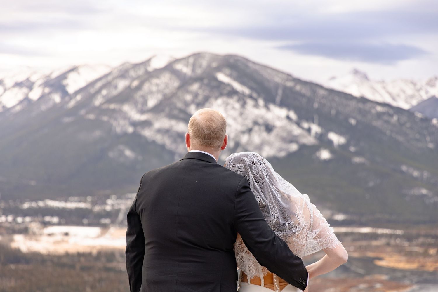 A couple stands on a mountain overlook with snow-capped peaks and dramatic clouds in the background.