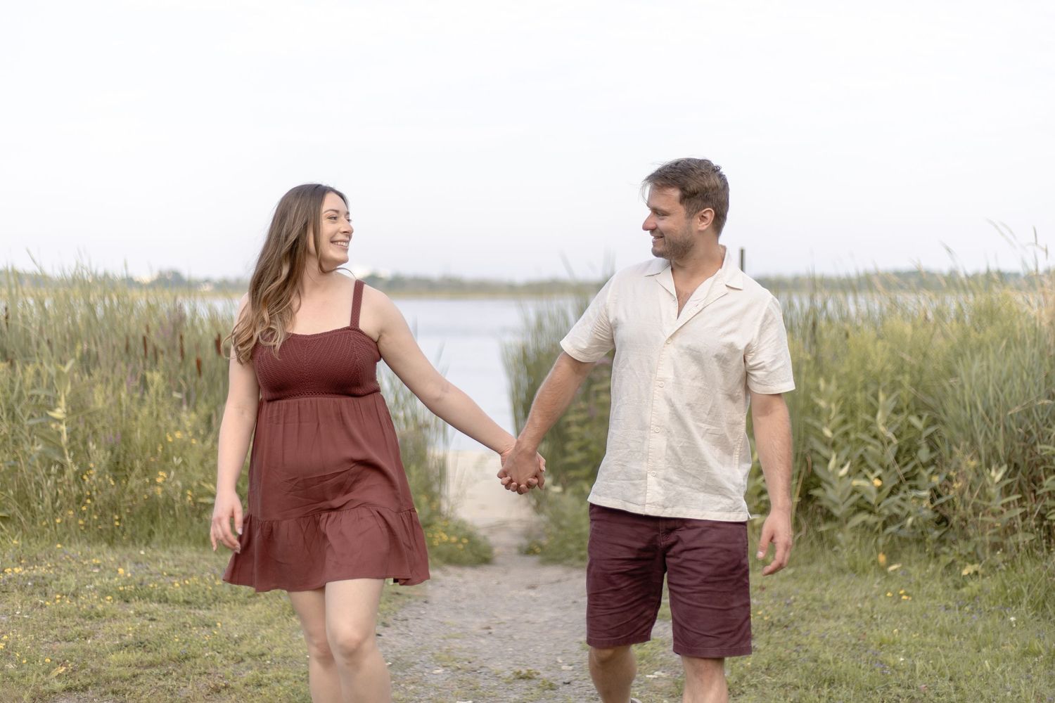 A couple shares joyful moments walking and laughing together on a scenic path through tall grass near the water.