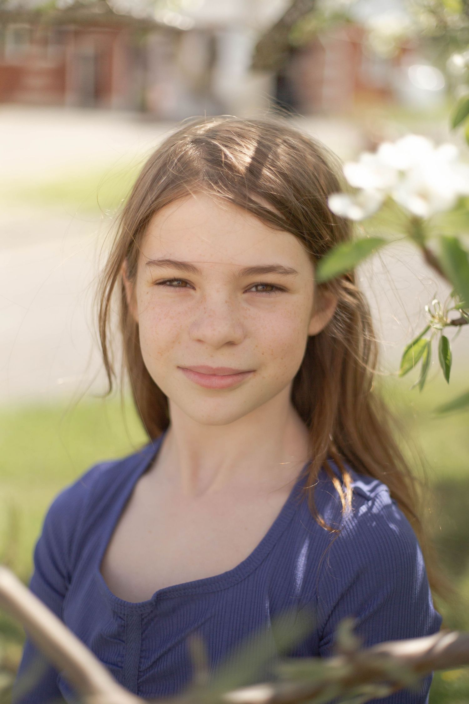 Young person in a navy blue top smiles warmly while standing outdoors near spring foliage on a sunny day.