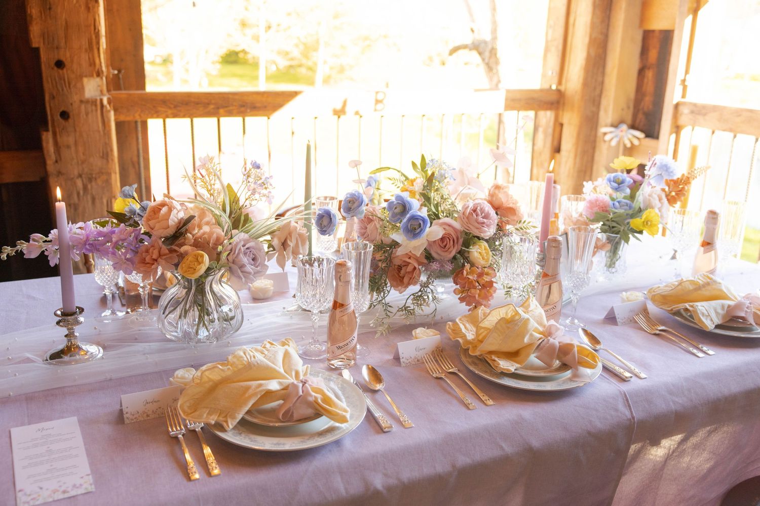 An elegant table setting with pastel floral centerpieces, crystal glasses, and gold napkins bathed in warm natural light.