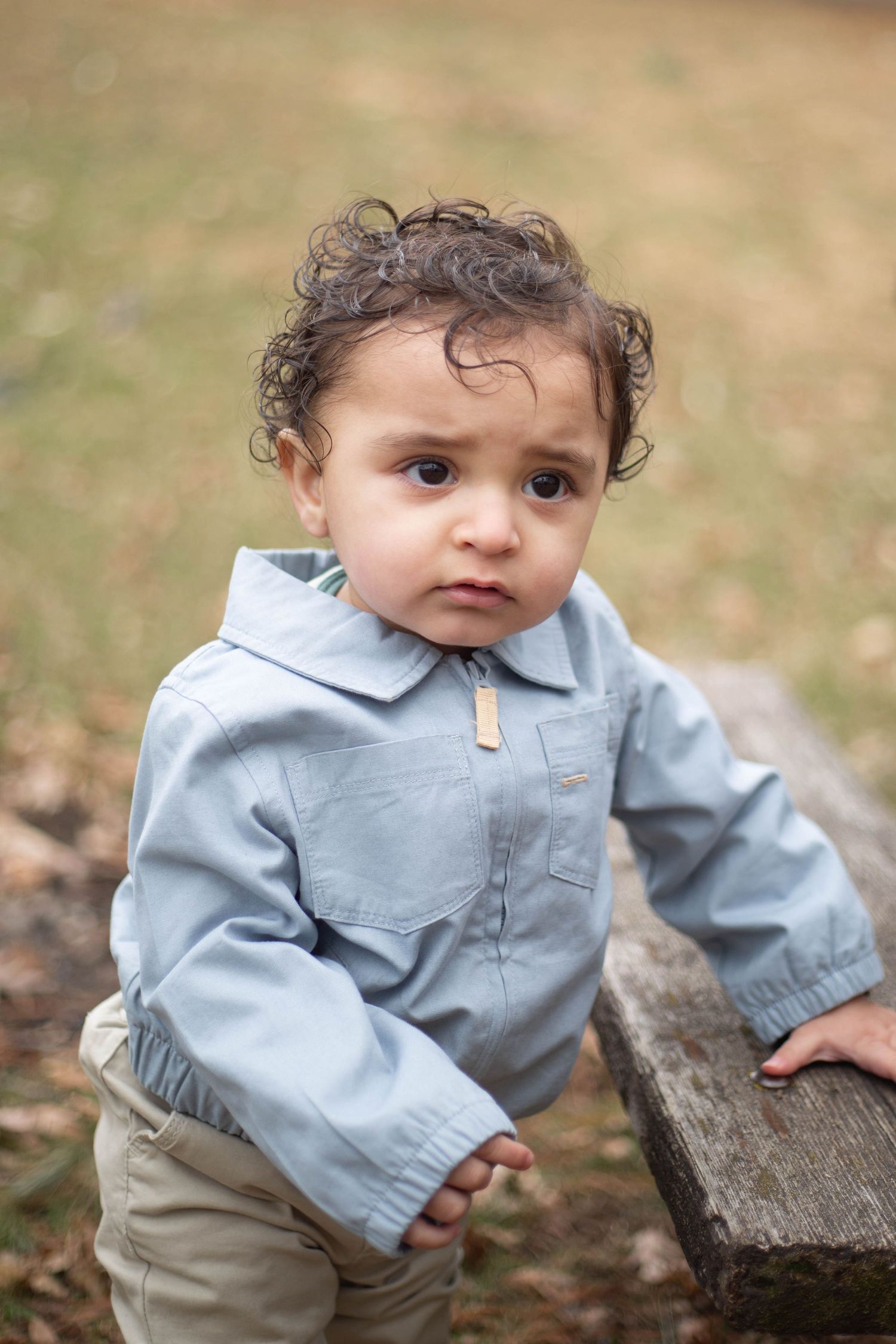 A toddler in a light blue jacket leans against a wooden bench in an outdoor setting.