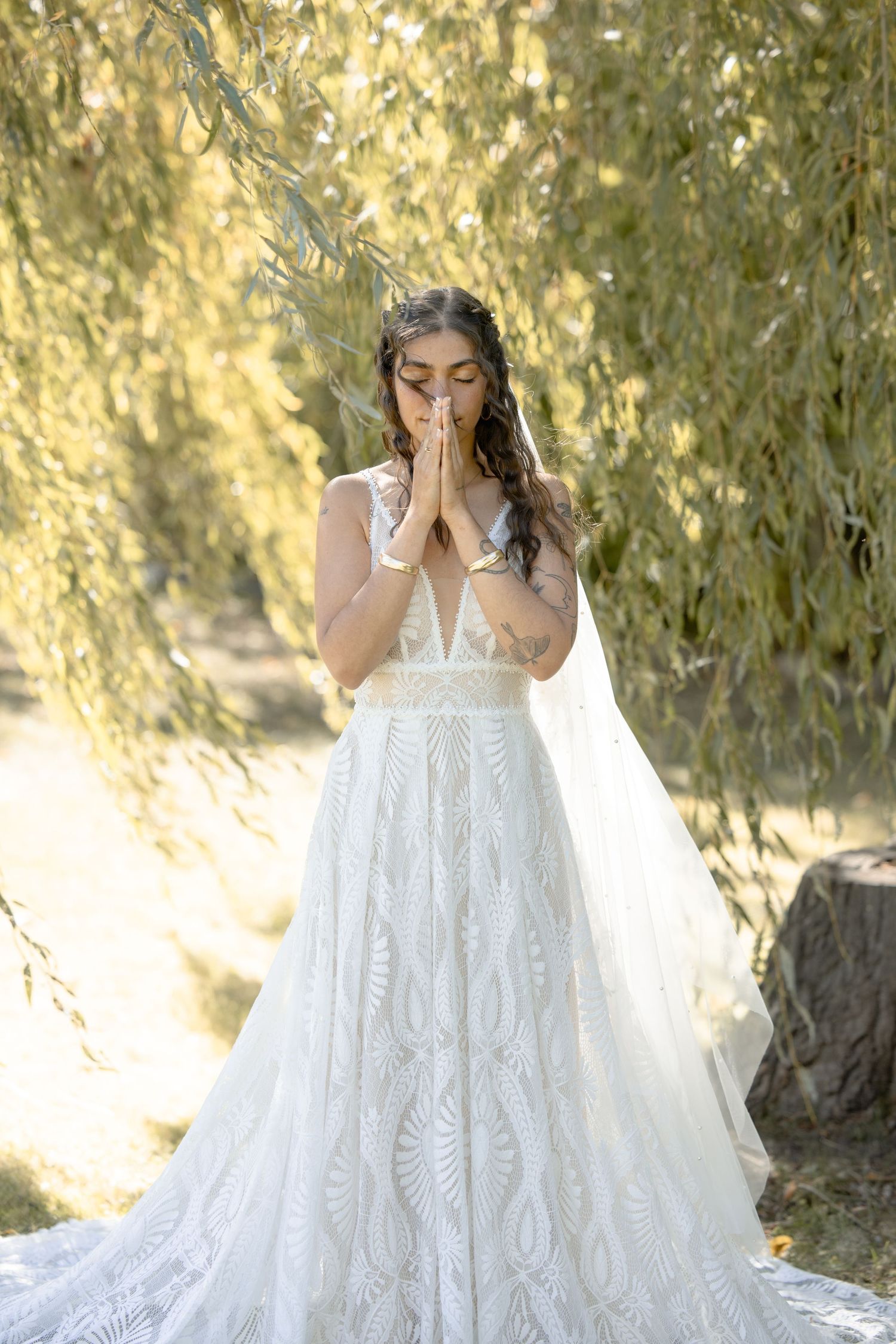 A bride in a flowing white lace gown poses emotionally under willow trees in golden sunlight.