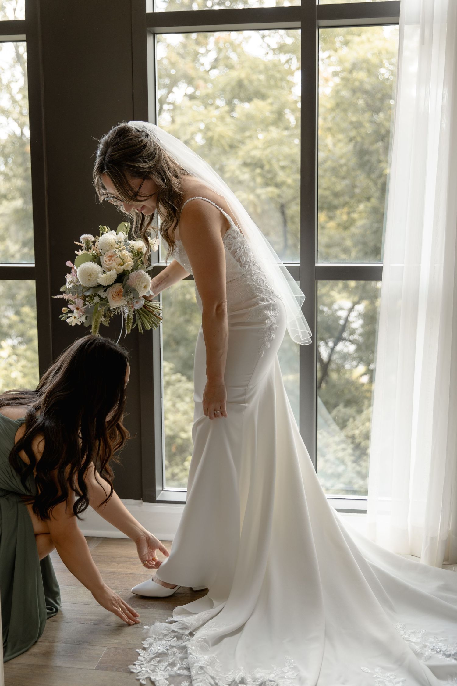 A bride in an elegant white lace wedding gown with long train gets help with her shoes near a bright window.