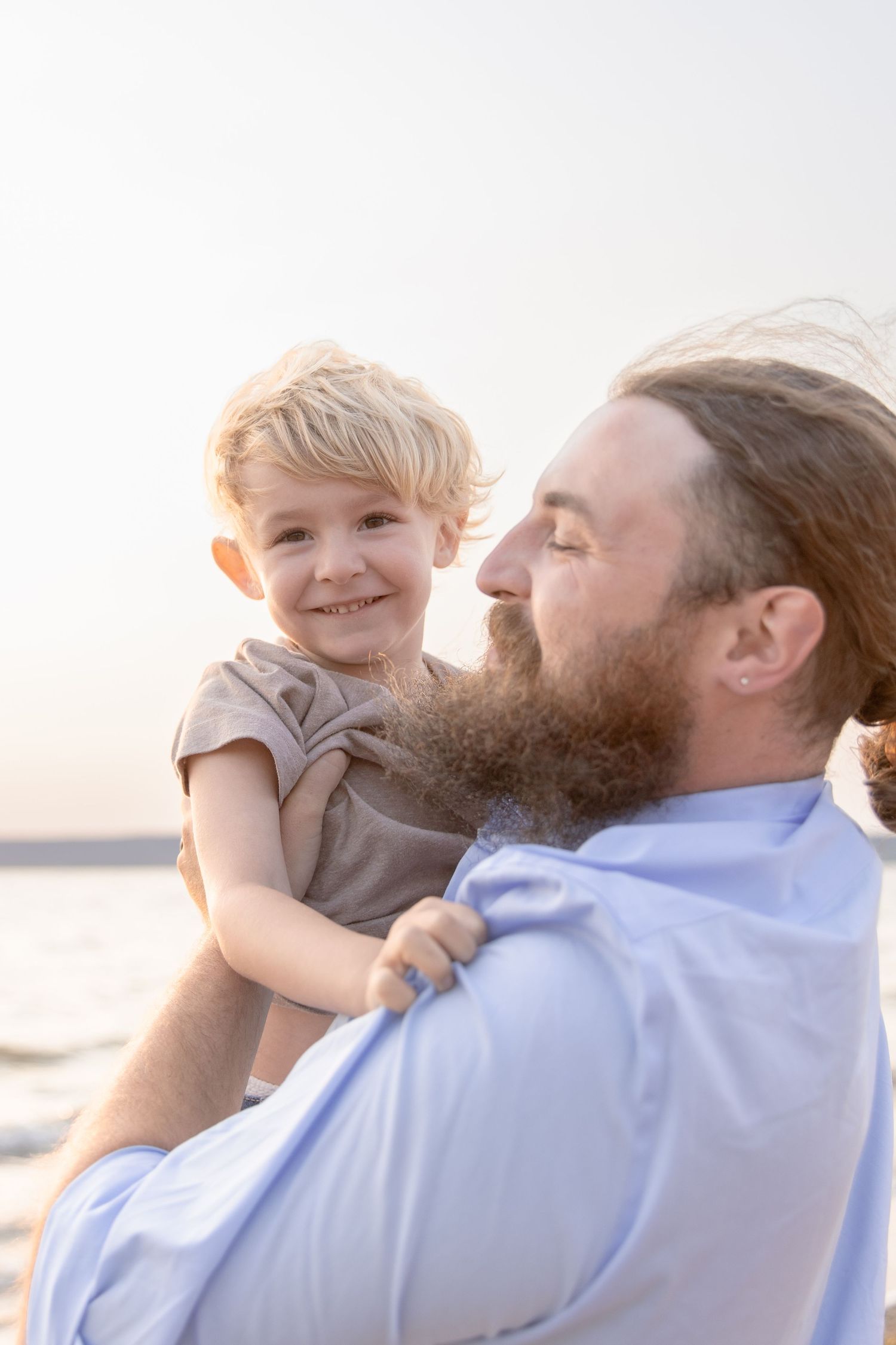 A bearded man in a blue shirt holds a young child while both enjoy a tender moment outdoors during sunset.