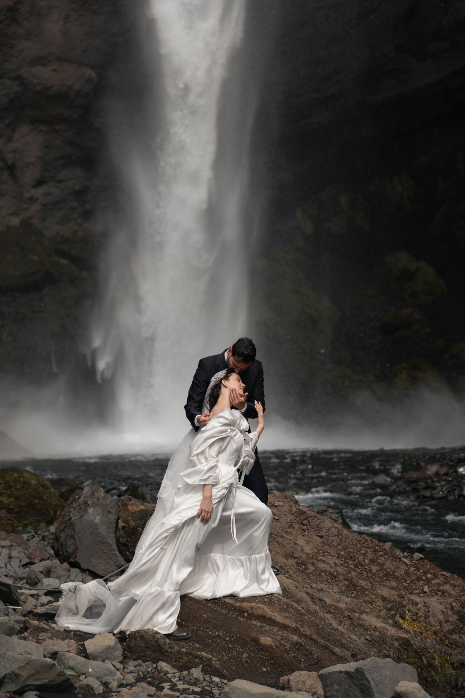 A bride and groom share a romantic kiss in front of a dramatic waterfall, her white gown flowing in the mist.