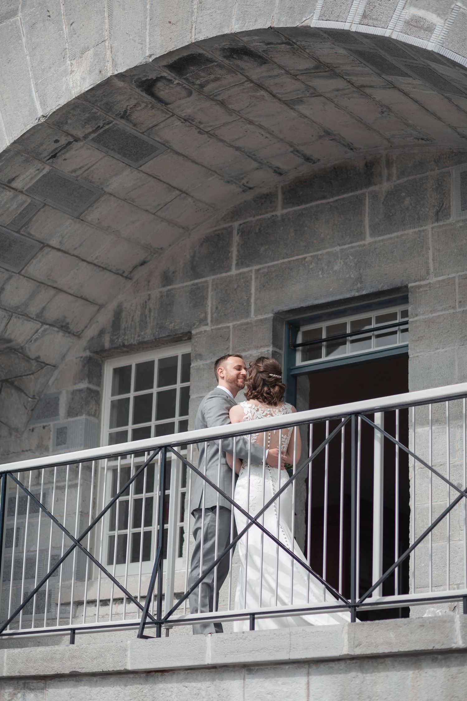 Bride and groom embrace on a stone balcony under an arch at their wedding venue.