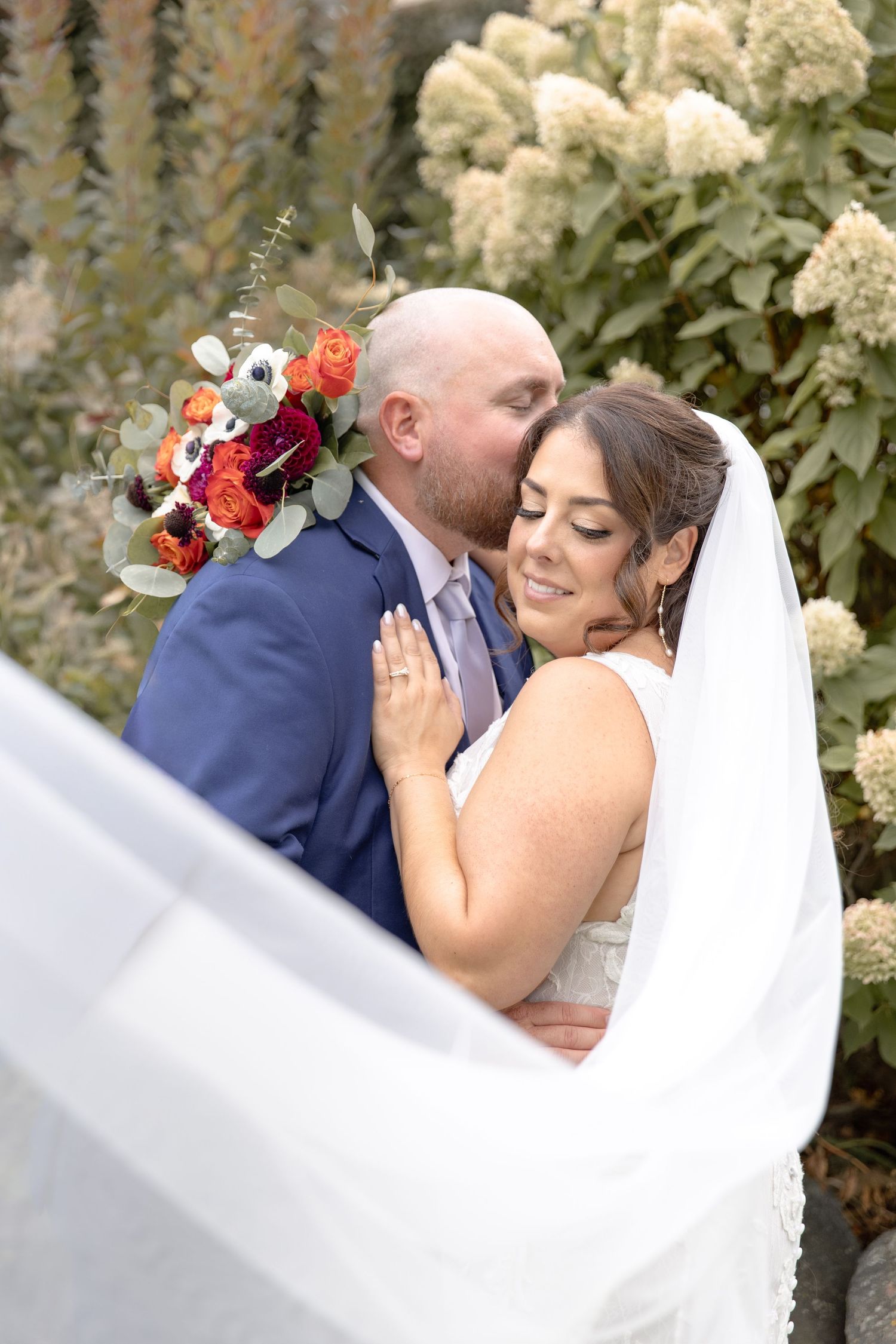 Romantic wedding couple embraces outdoors with flowing white veil and colorful floral bouquet against natural garden backdrop.