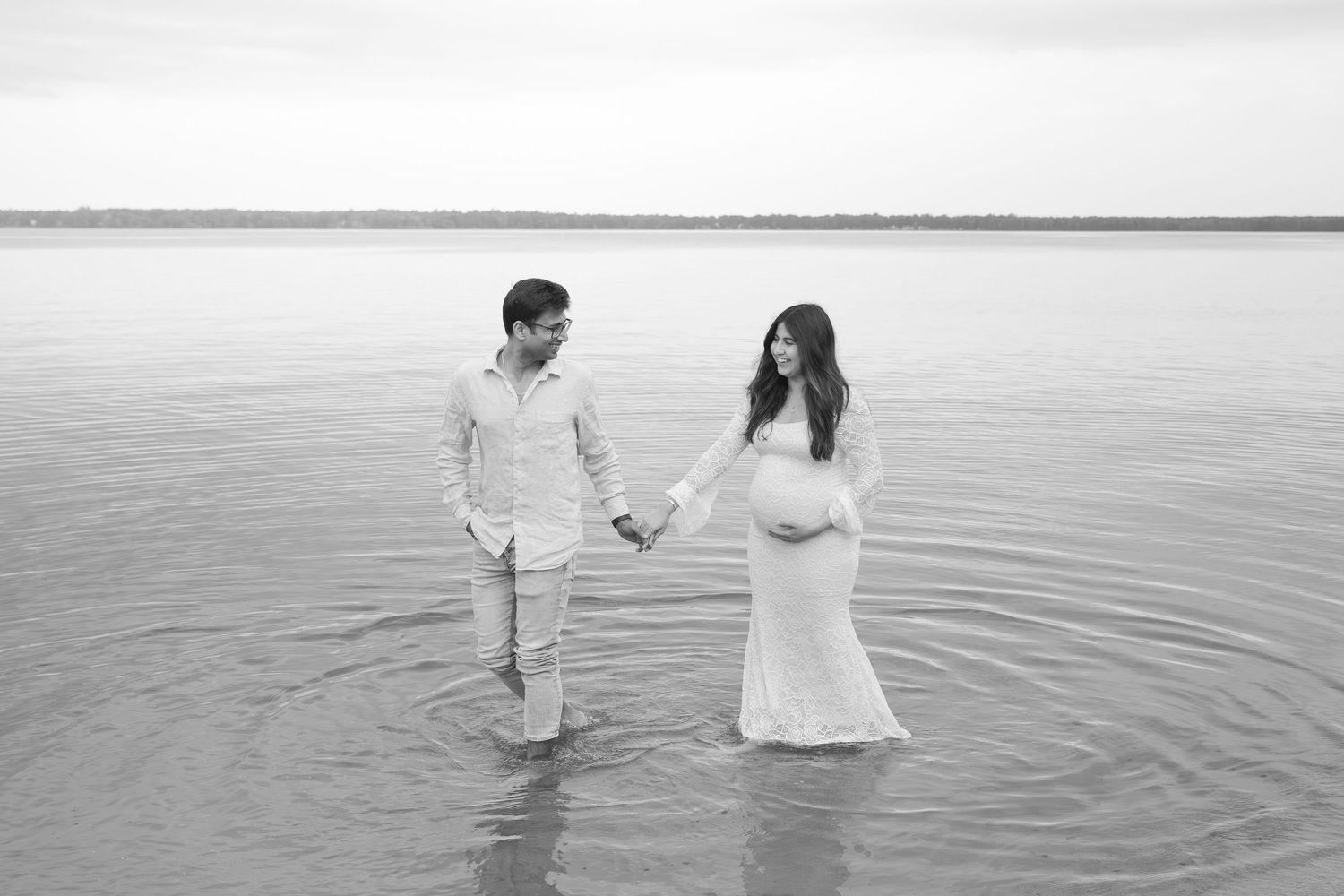 Two people in white outfits hold hands and walk in shallow ocean water during sunset creating a romantic scene.