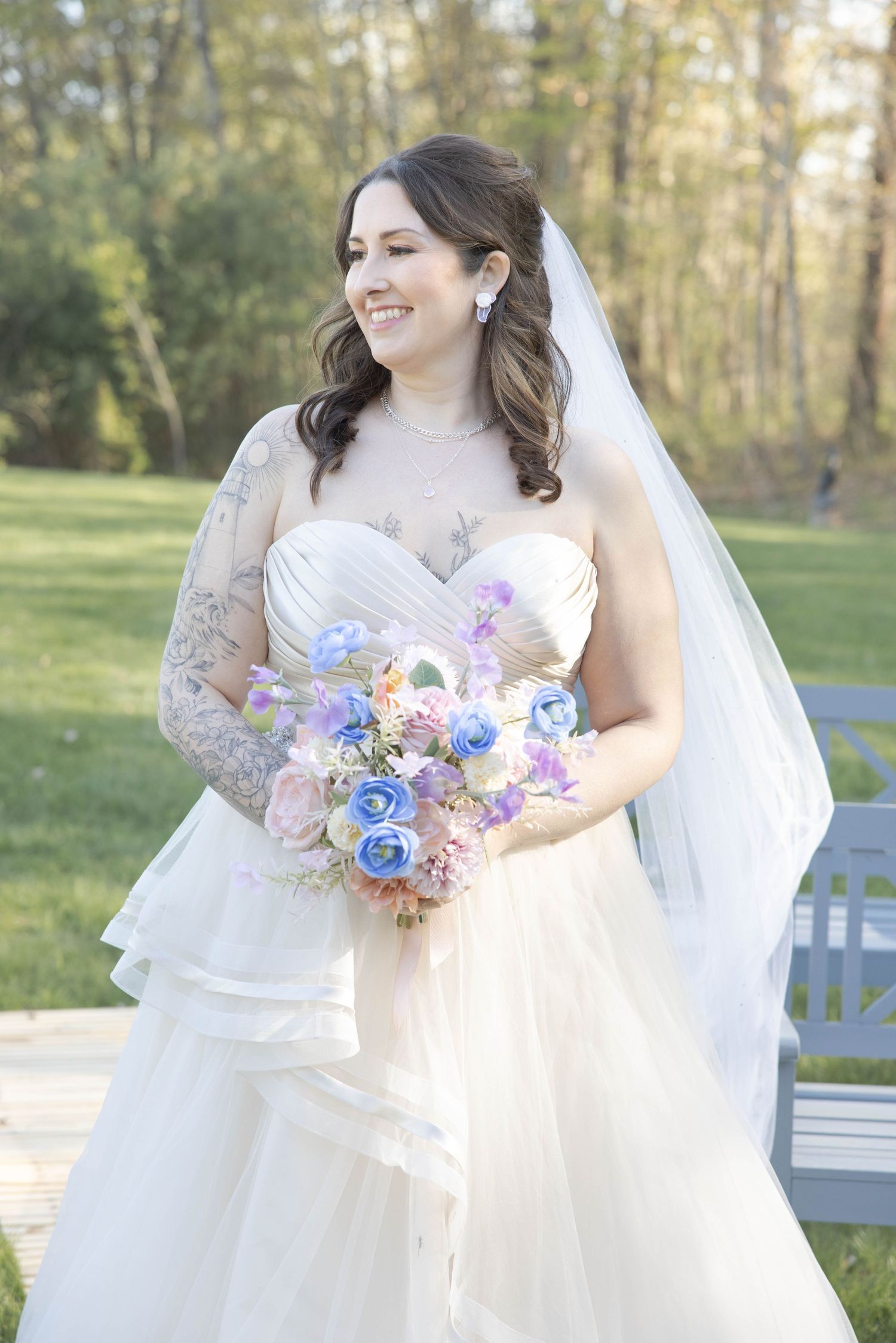 Bride in long-sleeved lace wedding gown holds pastel blue and purple bouquet while standing outdoors in a garden setting.