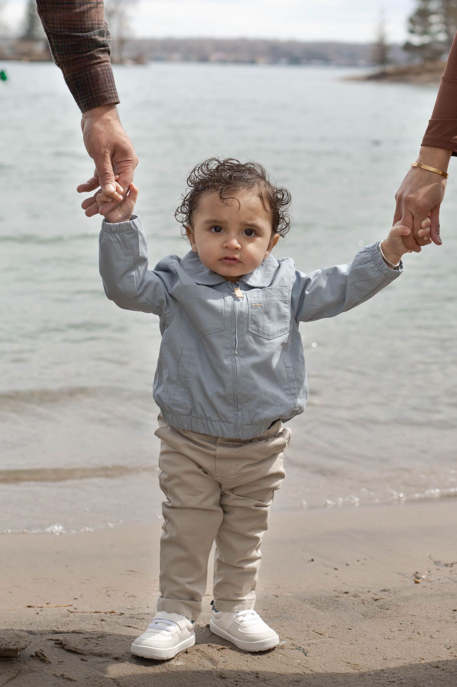 A small child in a light blue jacket and khaki pants holds onto adult hands while standing on a beach near the water.
