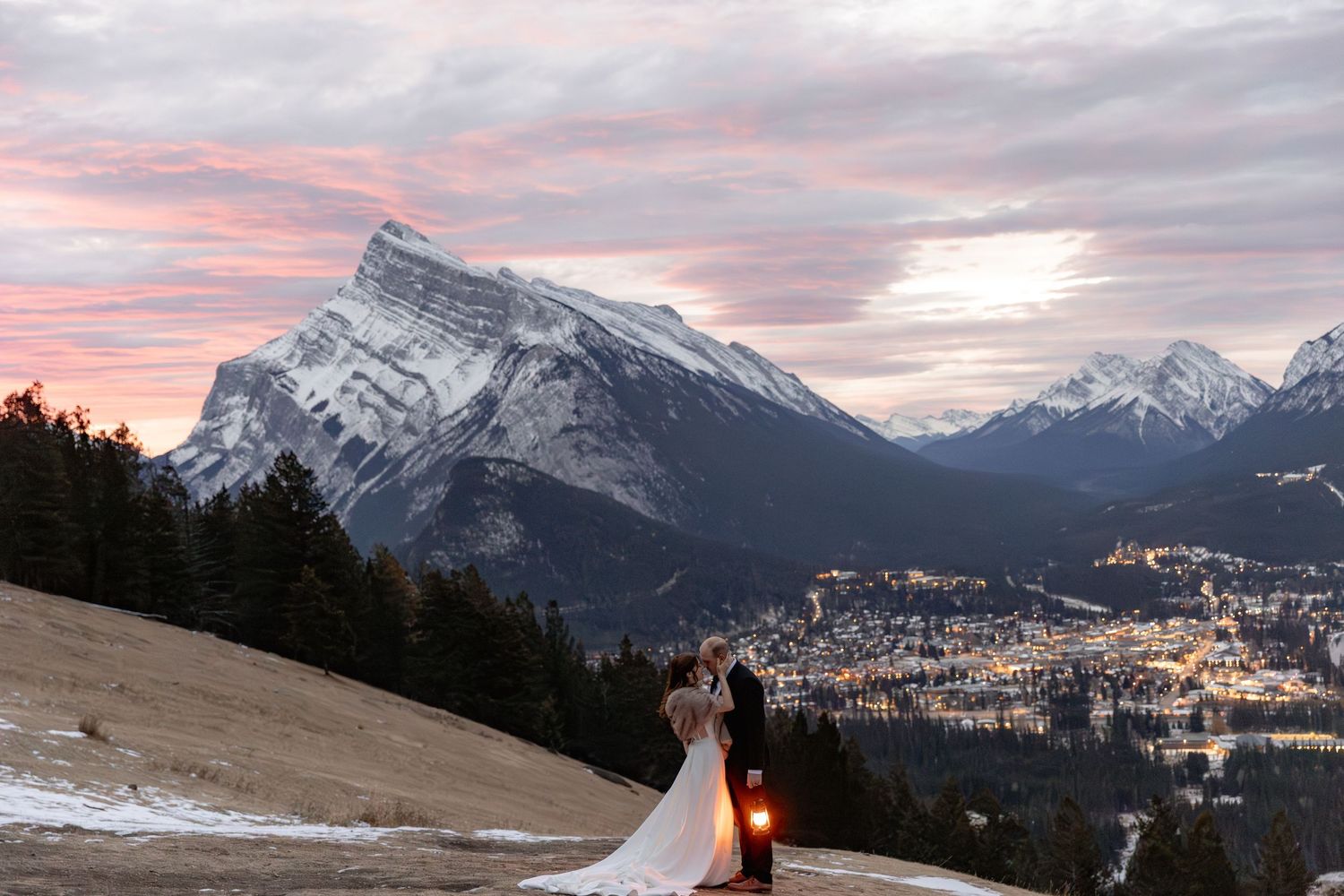 A snowy mountain landscape at sunset with a wedding couple silhouetted against the pink sky in Banff.
