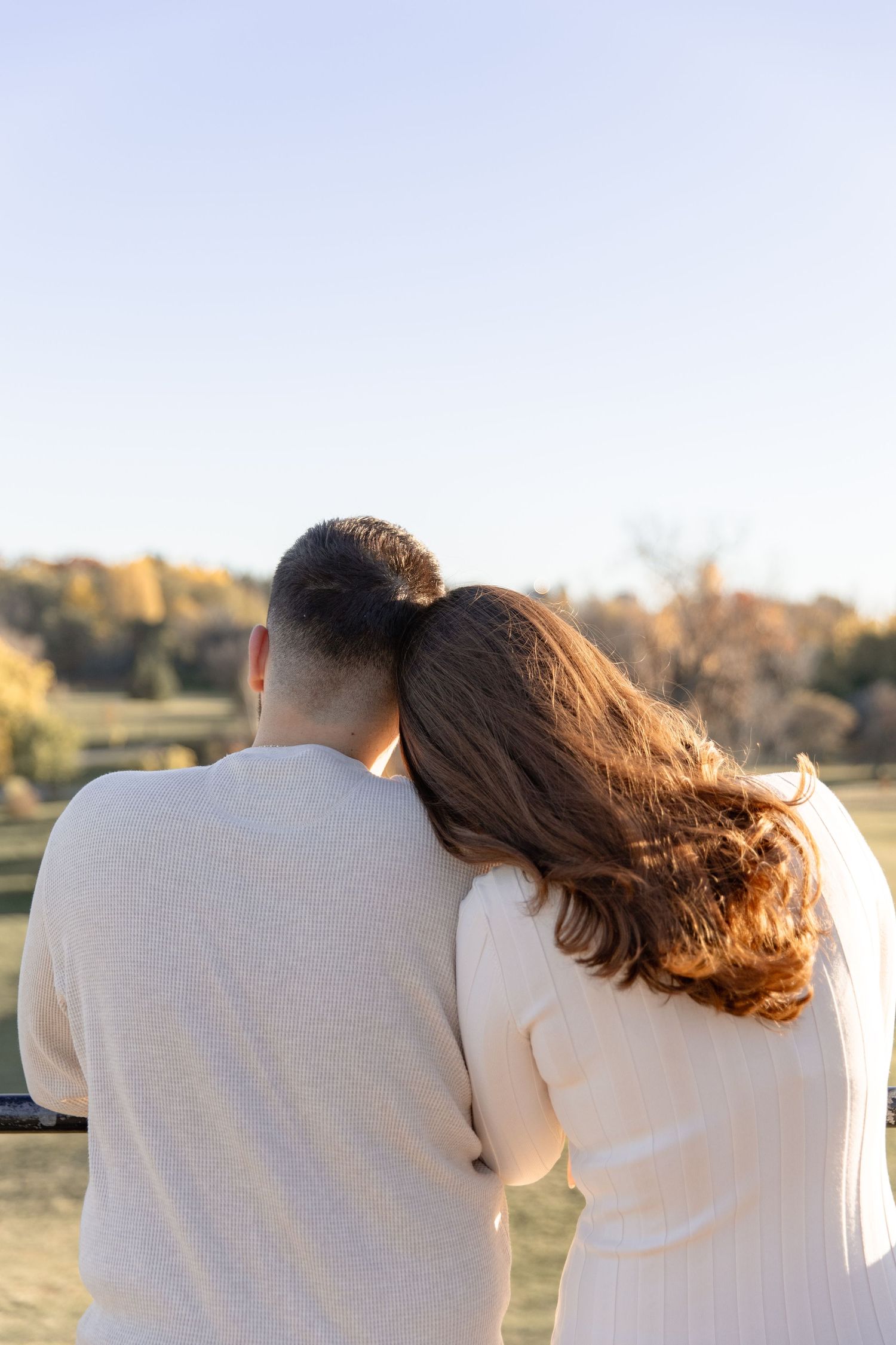 A couple in white sweaters sits together watching a sunset in a field during golden hour.