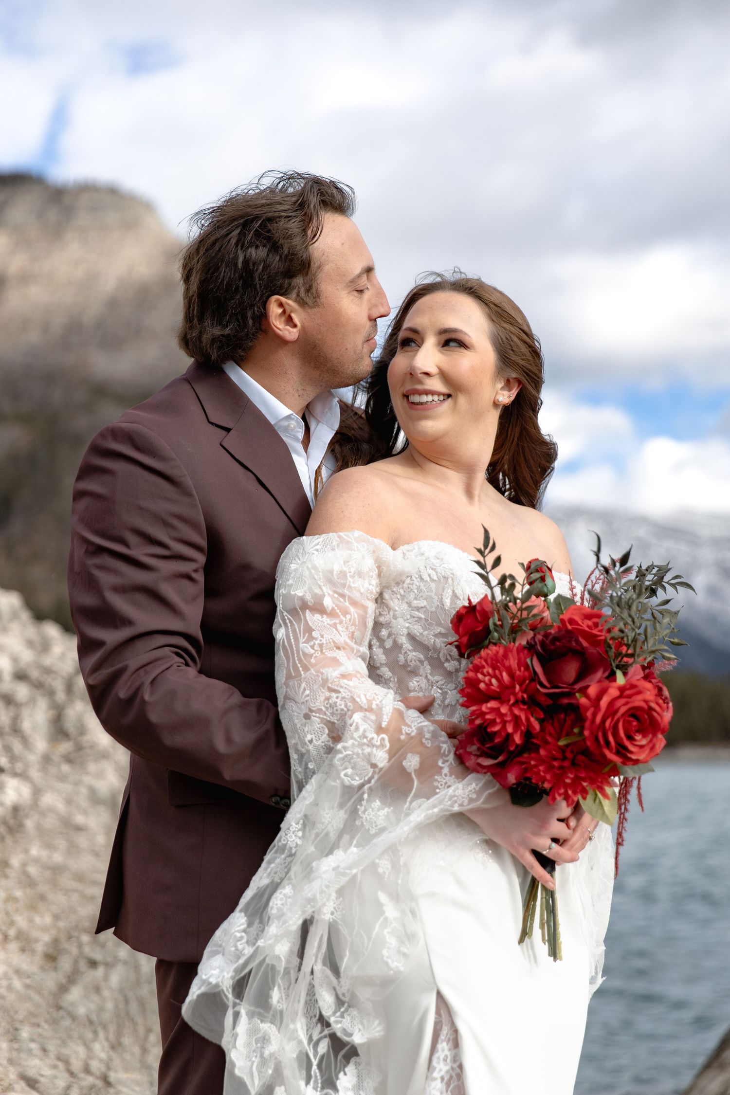 A couple in wedding attire embrace by the water, with the bride holding a vibrant red rose bouquet against a cloudy sky.