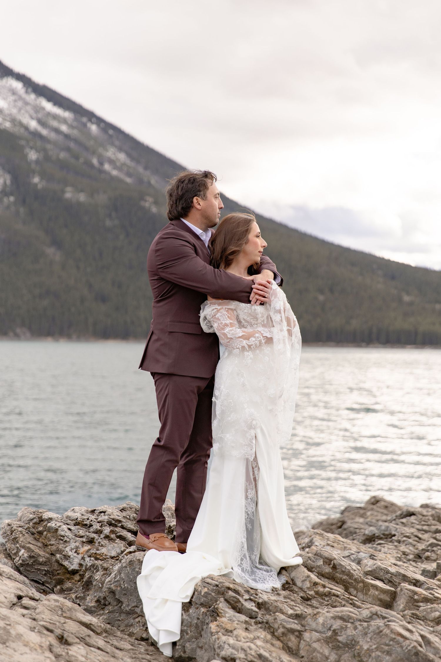 A couple in wedding attire embraces on rocky shores with mountains and lake in the background.