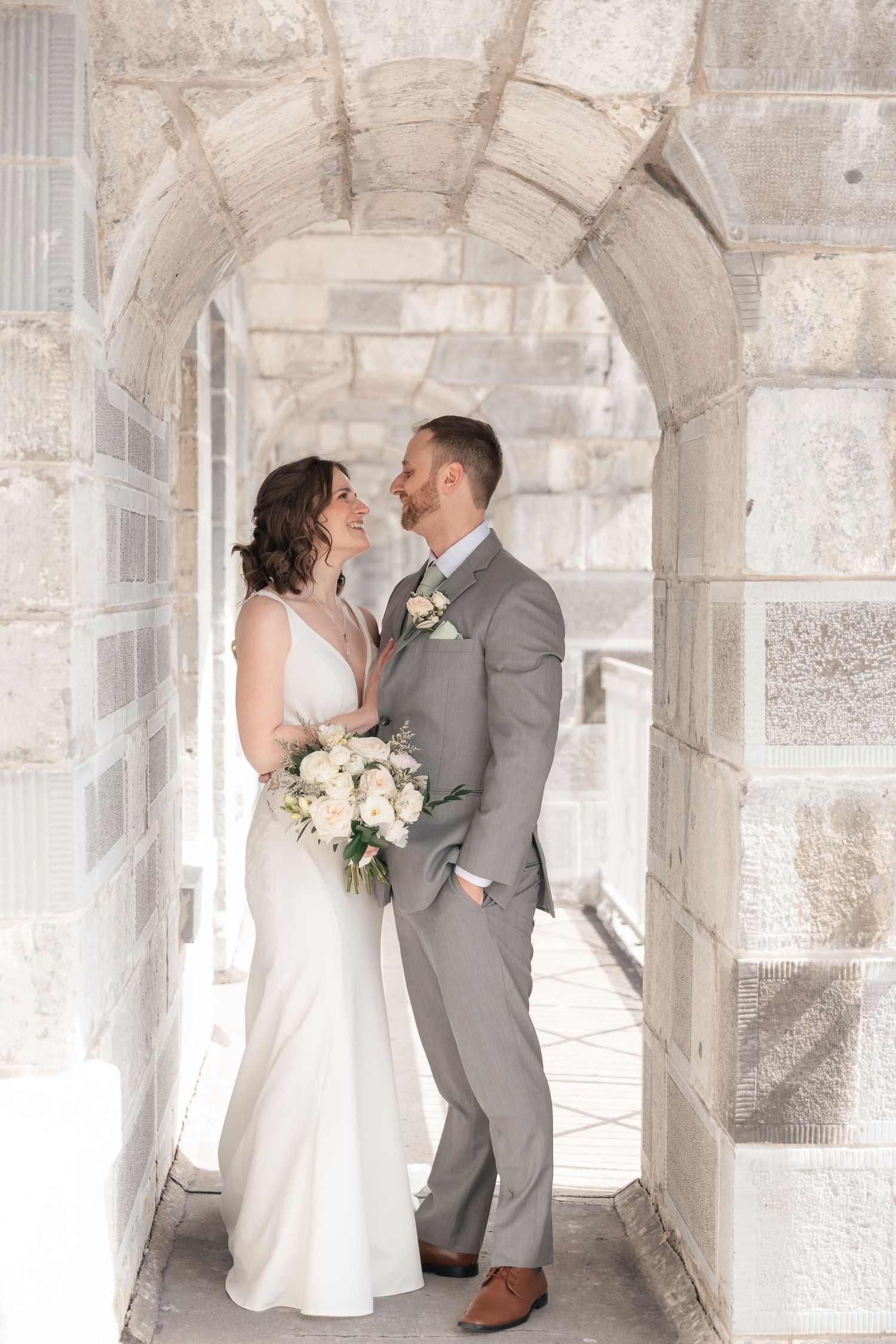 Bride and groom share a romantic moment under a stone archway, bride holding white bouquet, groom in gray suit.