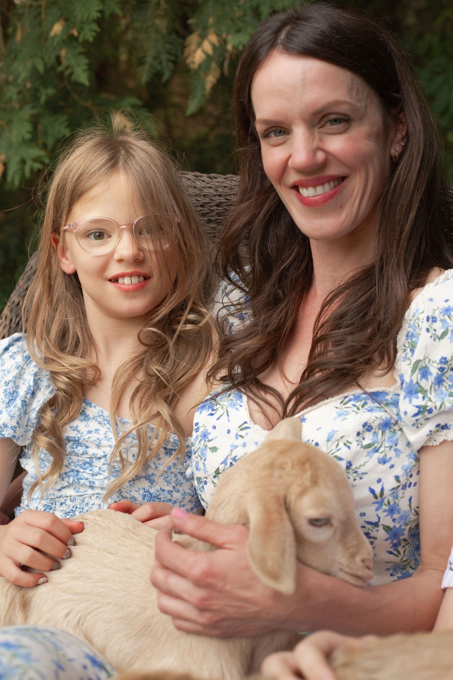 A pair in matching blue floral dresses hold and pet a cream-colored cat on their laps.