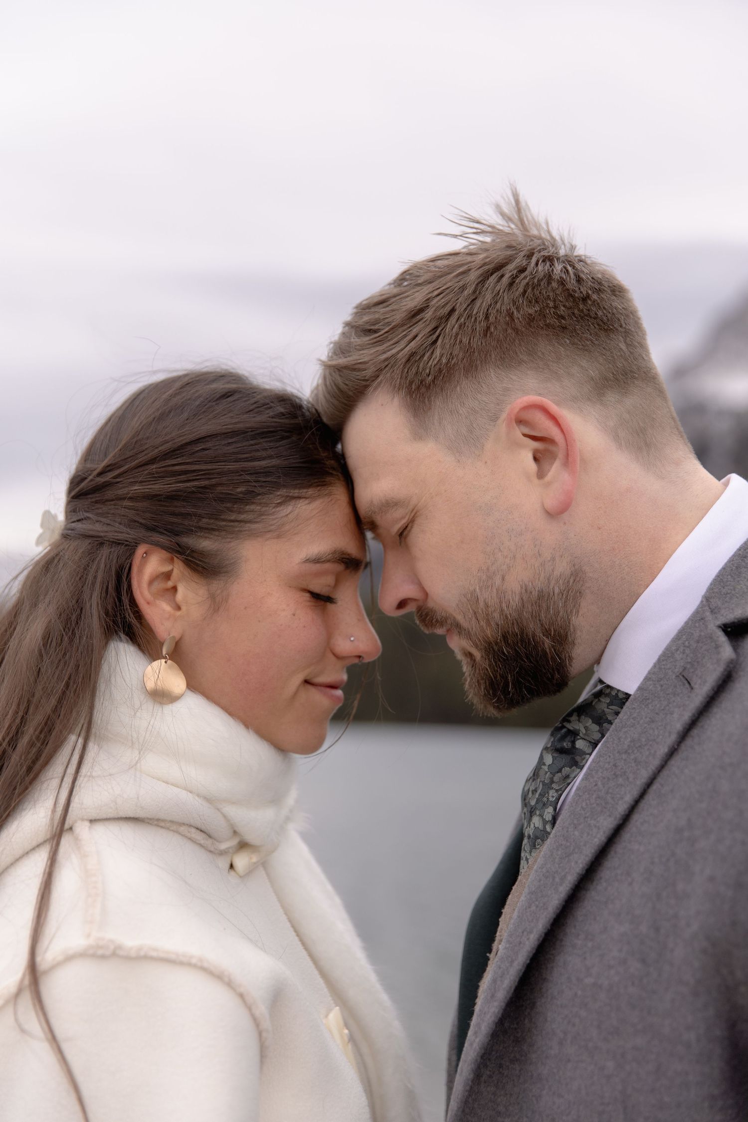 Romantic winter couple in white coat and grey suit share intimate moment against snowy mountain backdrop.