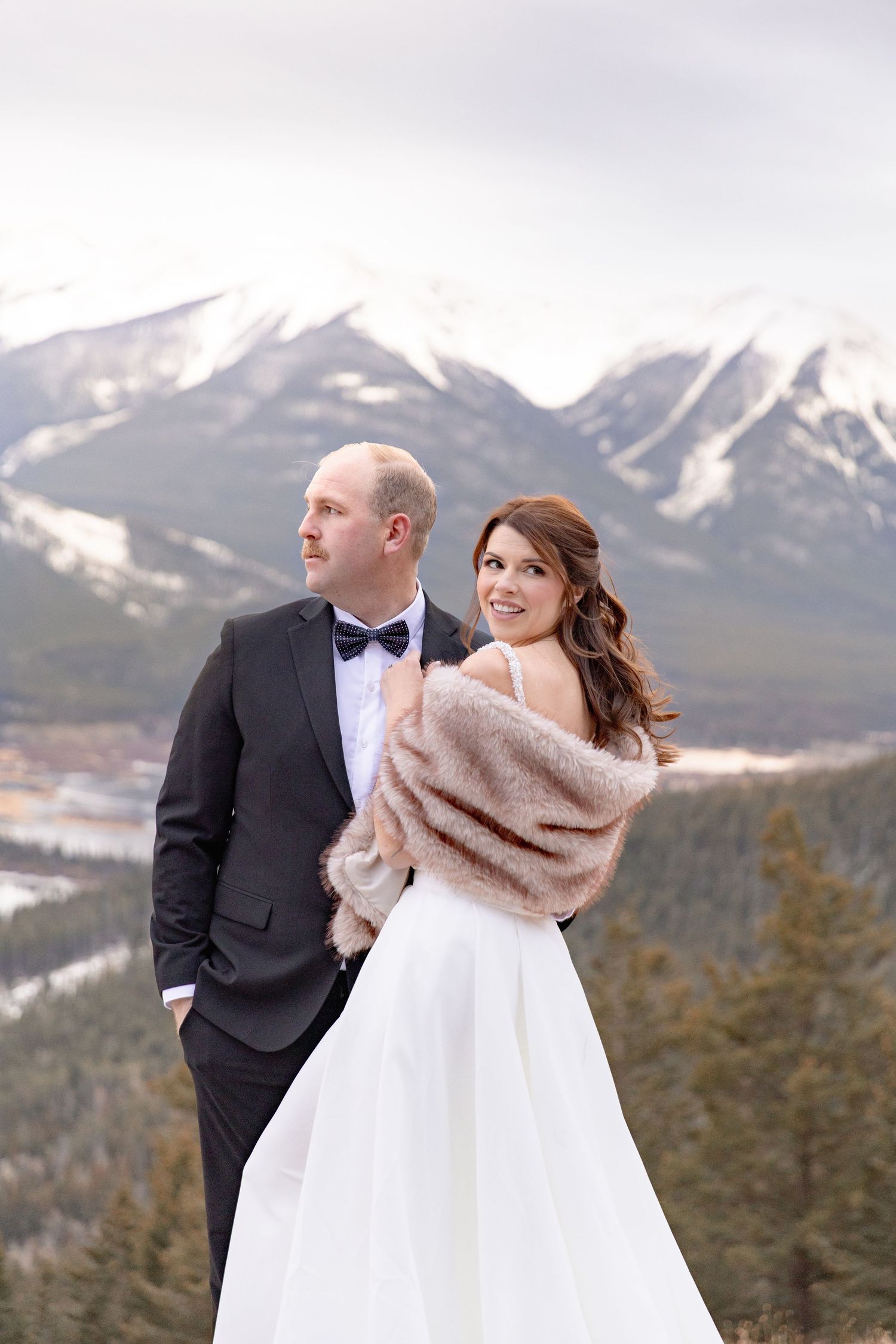 A couple in wedding attire poses together against a snowy mountain backdrop in winter.