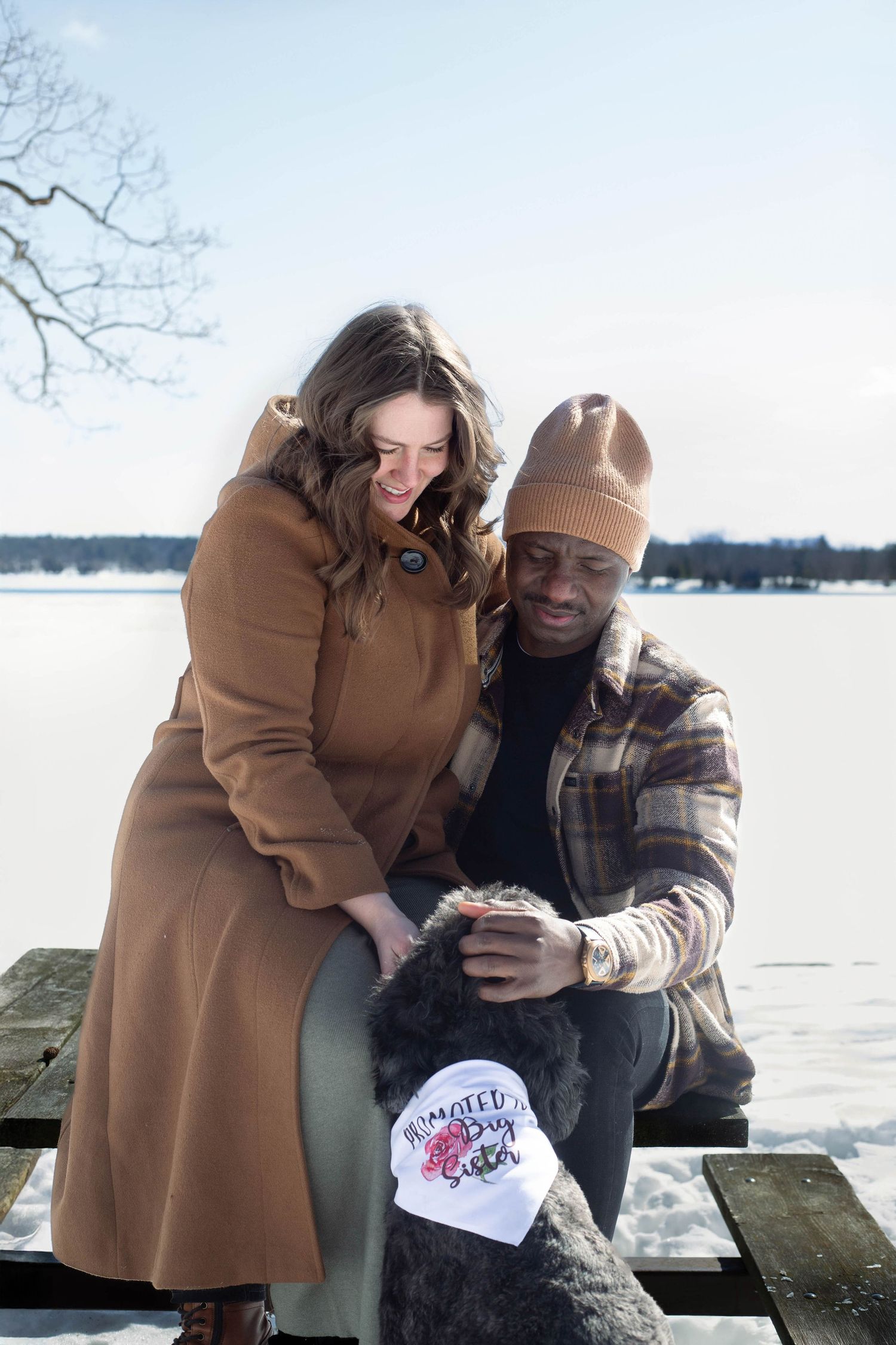 A couple sits with their black dog on a dock overlooking a frozen lake on a winter day. The dog wears a 