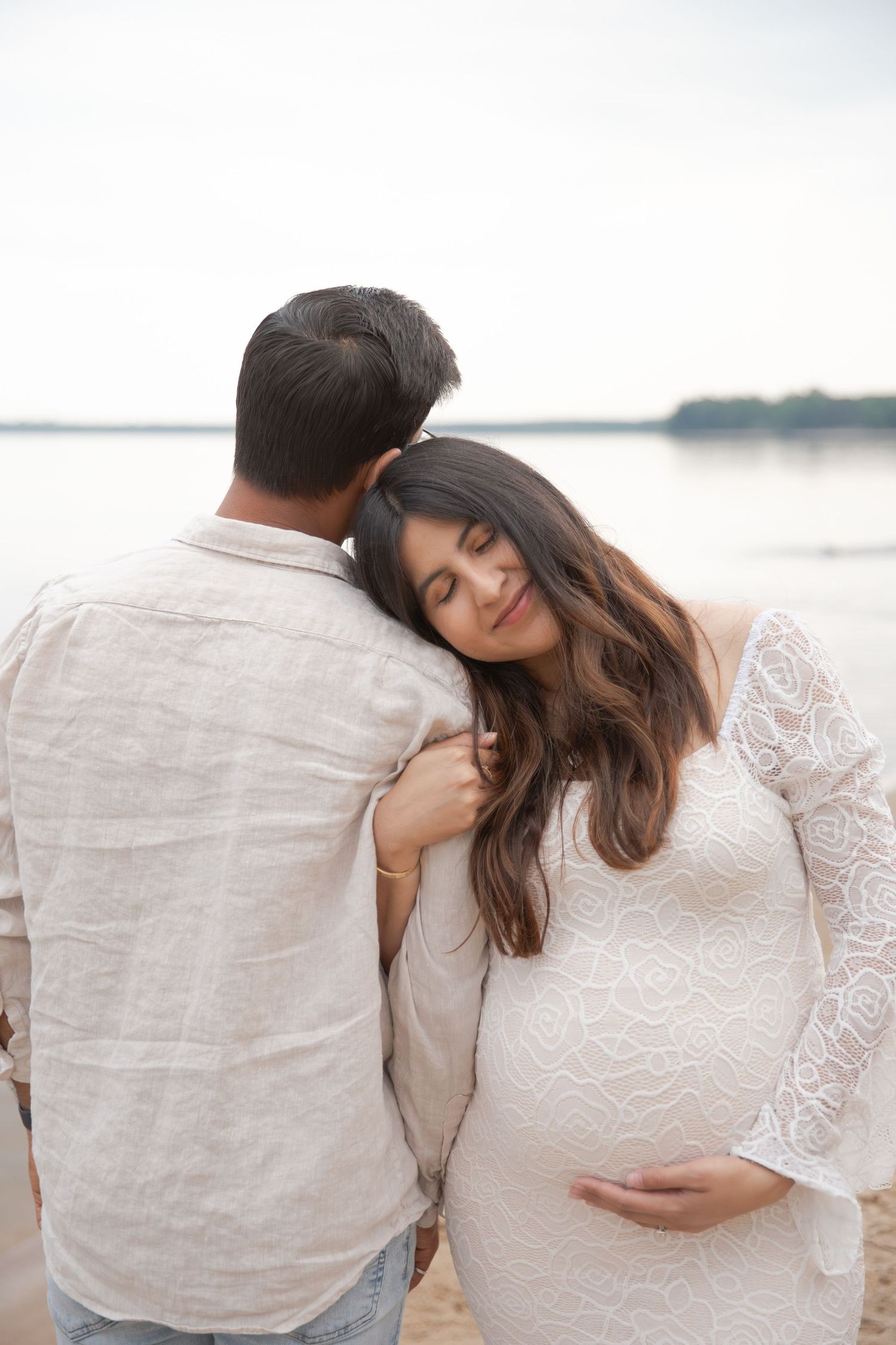Expecting couple in white clothing embrace tenderly on a beach at sunset while cradling the pregnant belly.