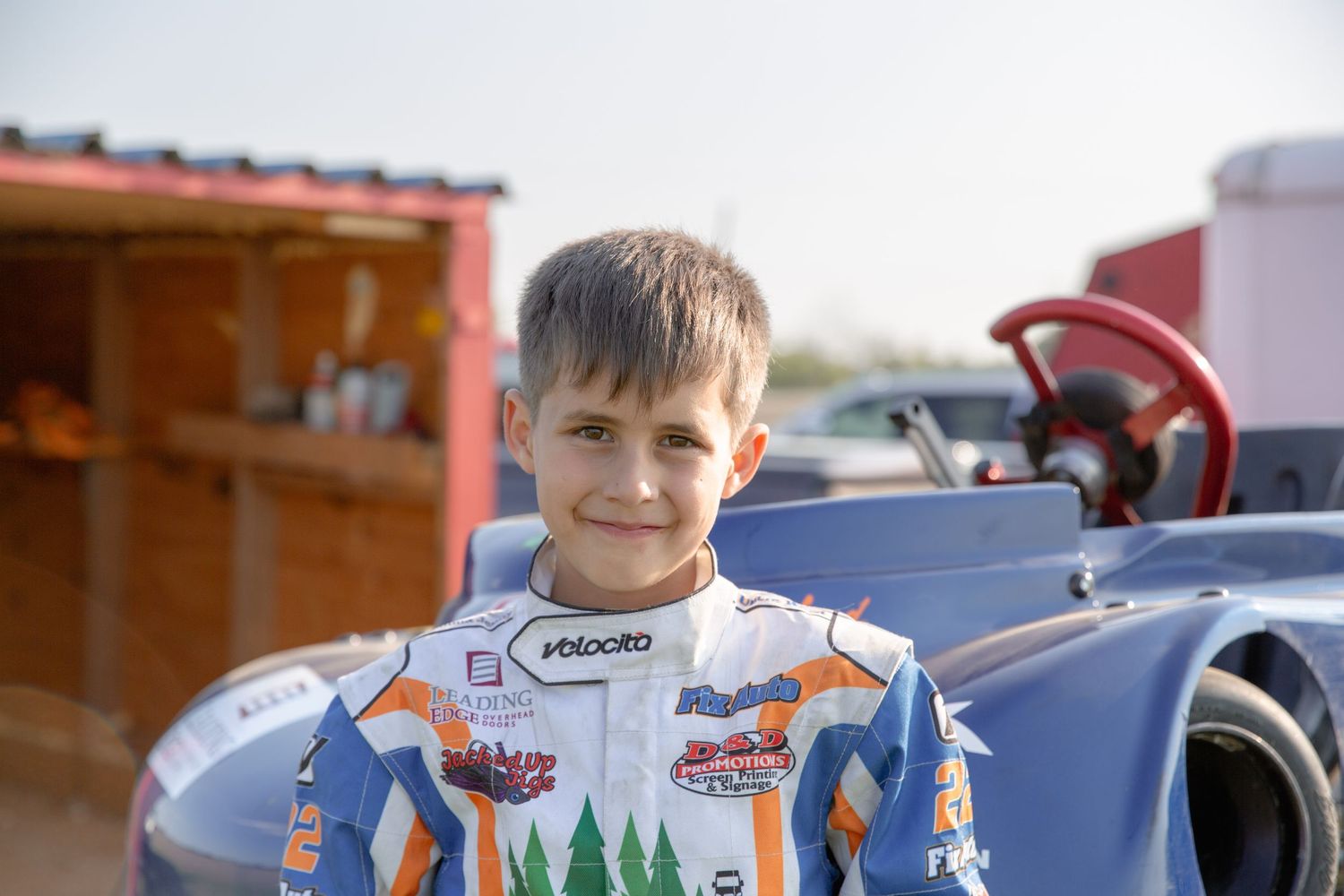 A person in white race gear and helmet sits in the cockpit of a motorsport vehicle with orange and red colors.